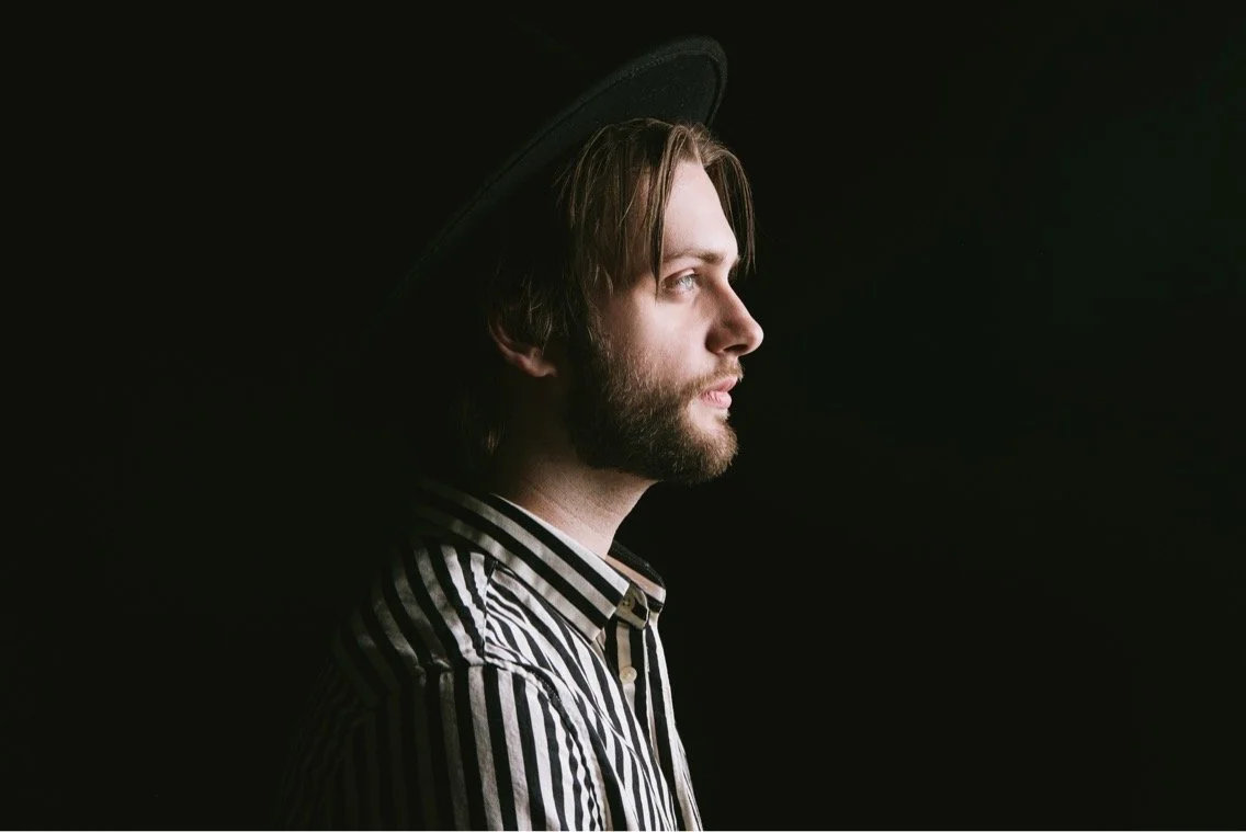 Side profile of a young man with a beard, wearing a striped shirt and a black hat, against a dark background.