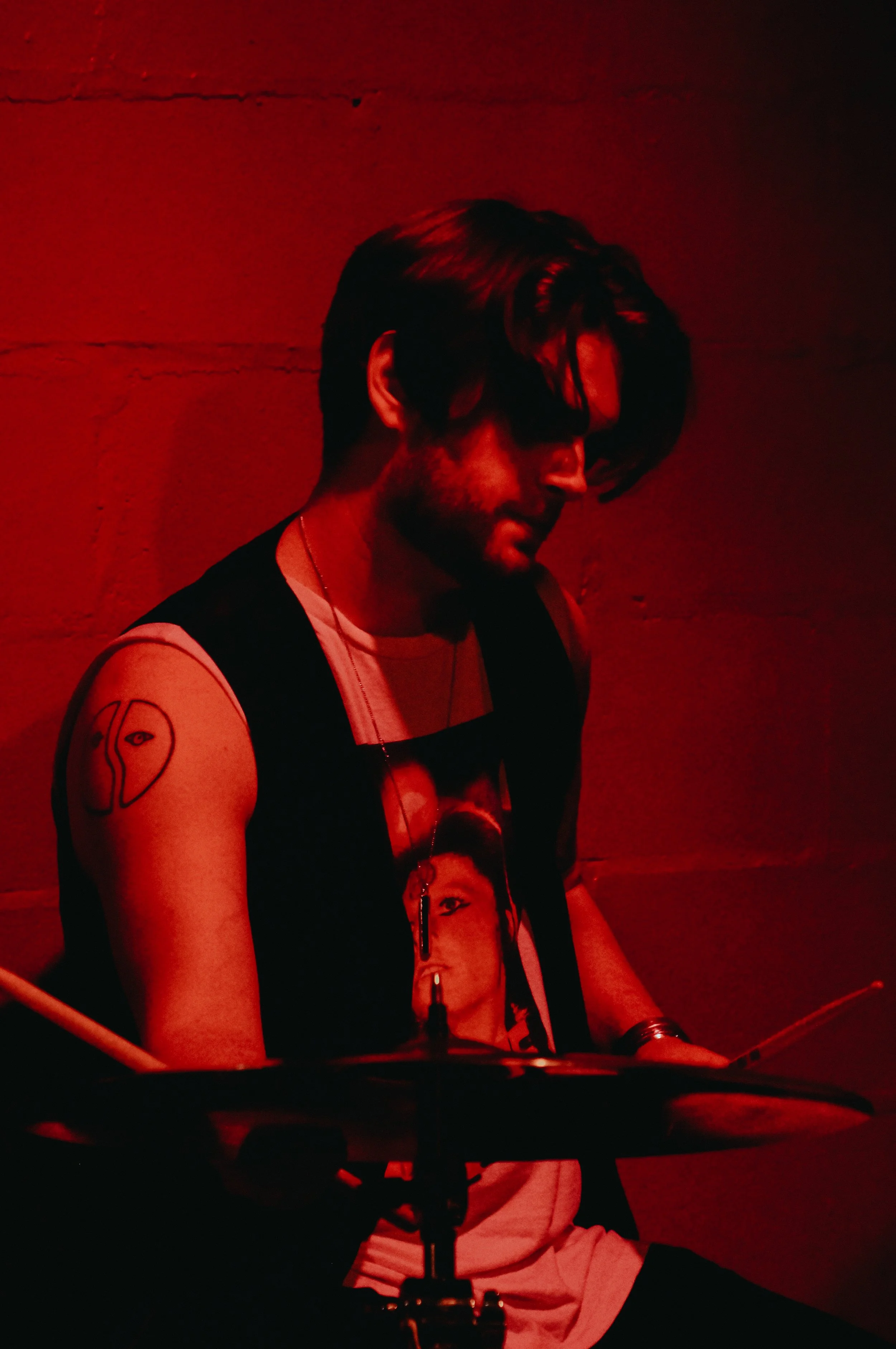 A person with shoulder-length hair and a beard, wearing a sleeveless shirt with a graphic, is playing drums in a room with red lighting, sitting behind a drum kit.