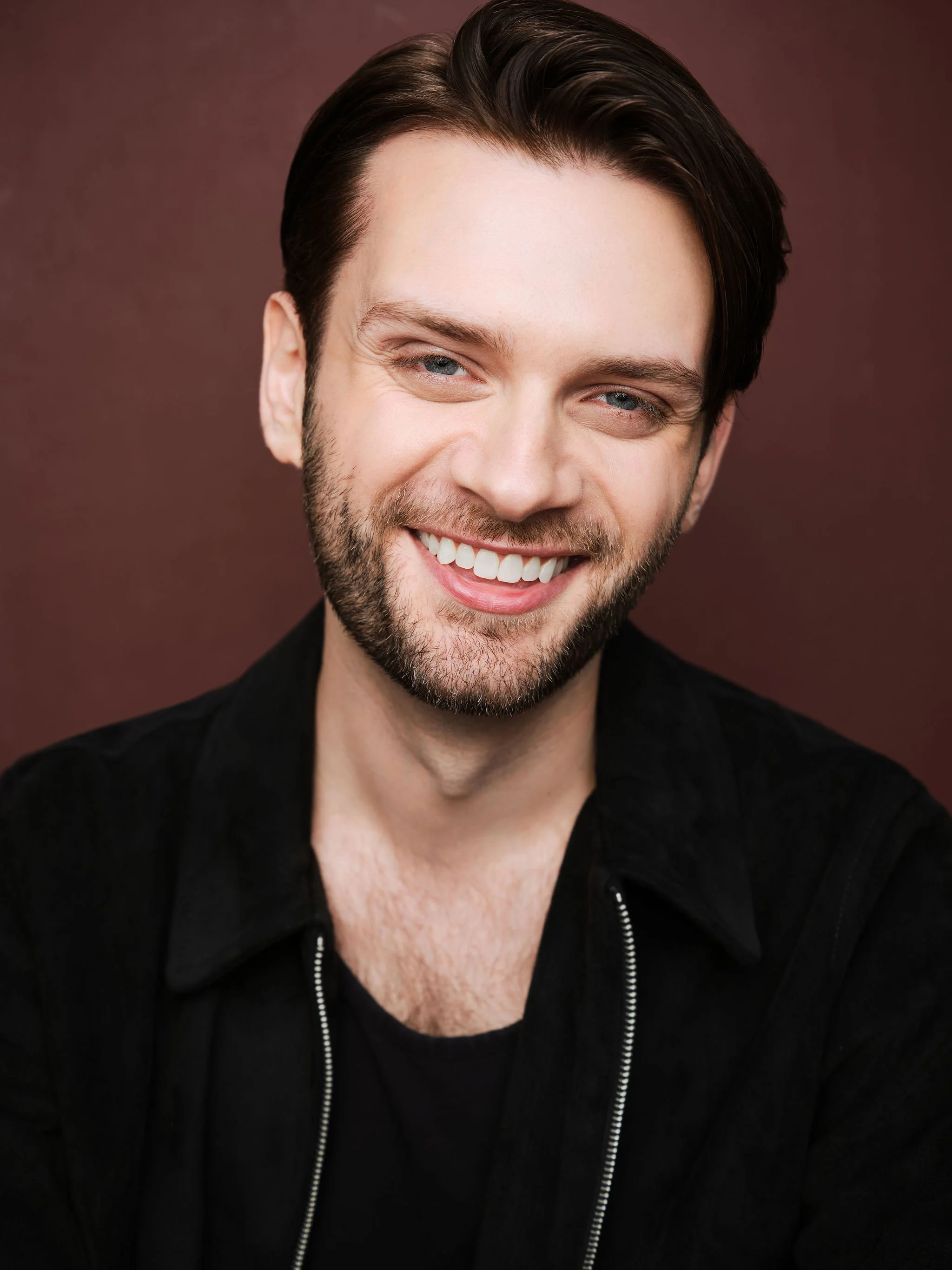 A young man with dark hair, blue eyes, and a beard, smiling in front of a plain brown background.