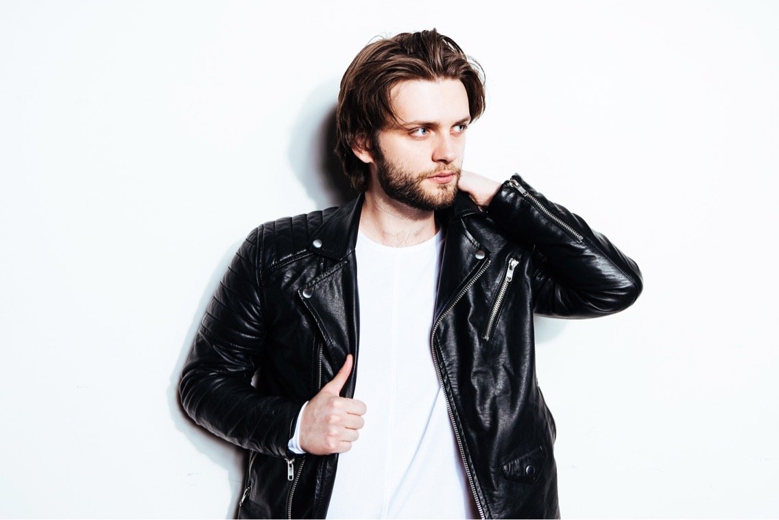A young man with brown hair and a beard wearing a black leather jacket and white T-shirt, standing against a white background, looking to the right, with one hand touching his neck and the other holding the jacket.