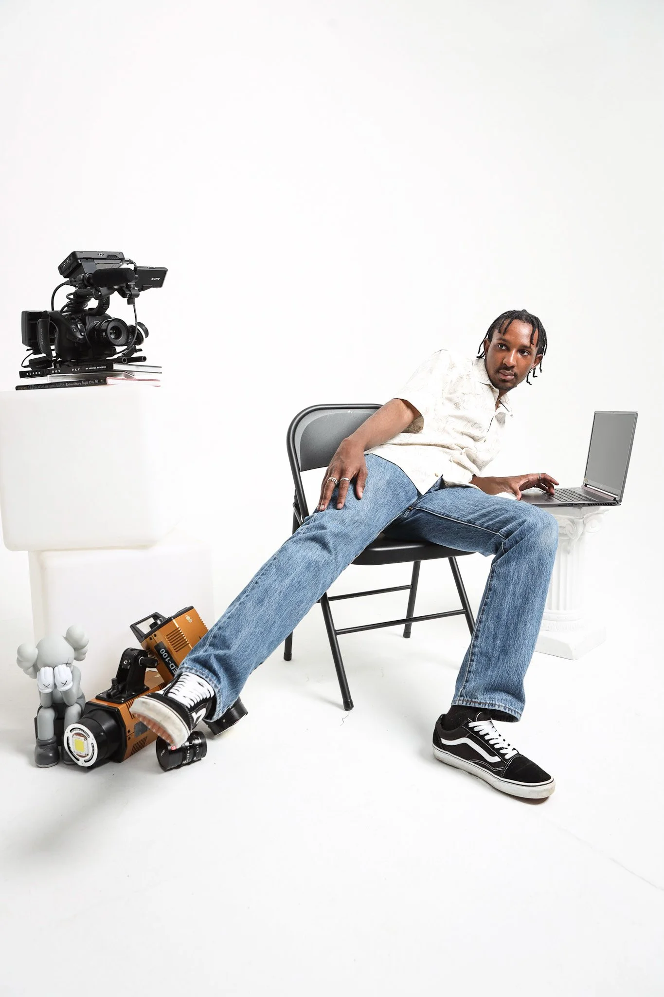 A man sitting on a black chair with his legs extended and resting on the floor, leaning to the side against the chair's armrest. He is wearing a white shirt, blue jeans, and black sneakers with white laces. There is a laptop on a white pedestal to hi