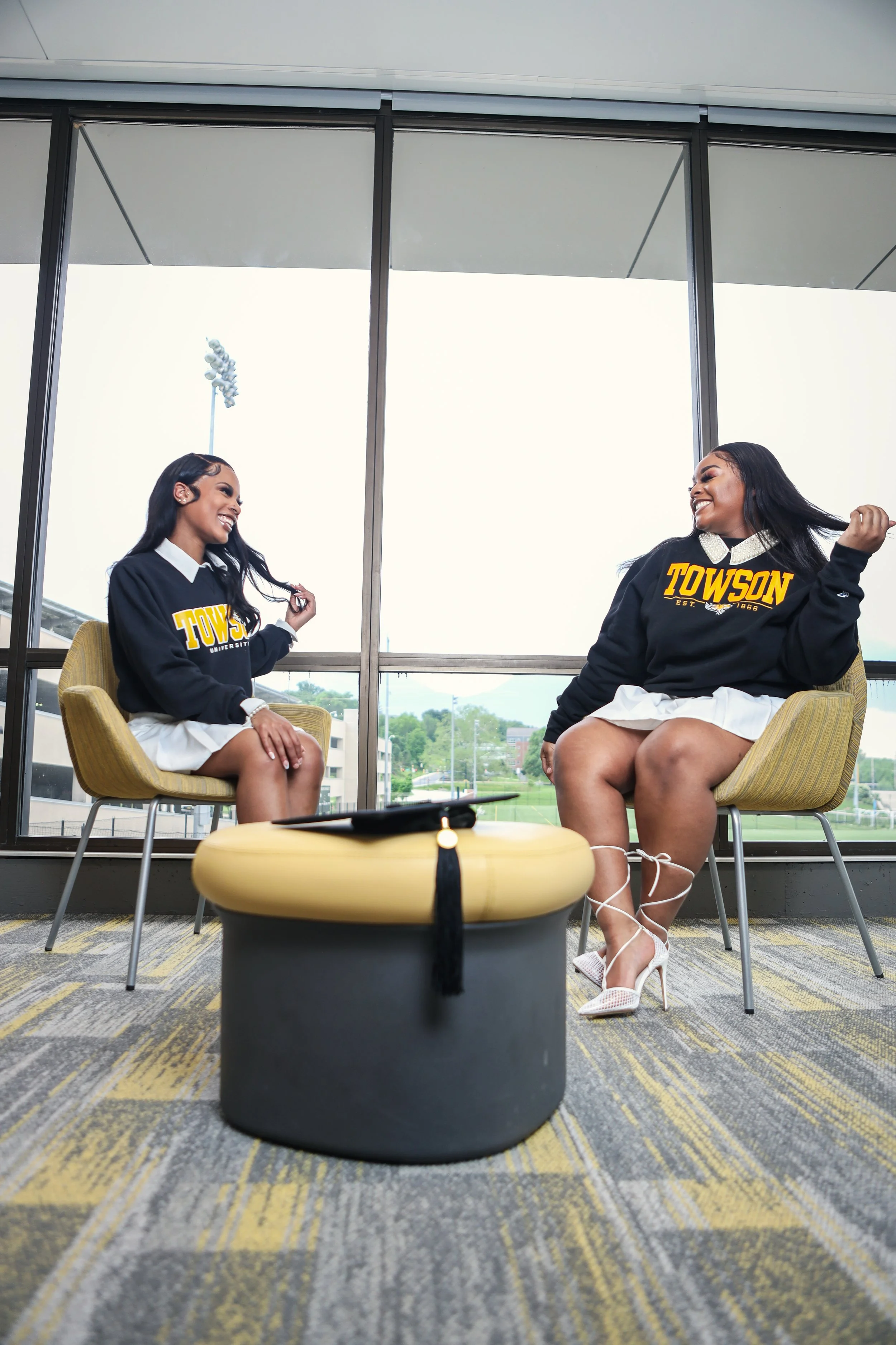 Two women in graduation robes sitting in chairs, smiling at each other, with a graduation cap and tassel on a table in front of them, inside a room with large windows showing trees and a field outside.