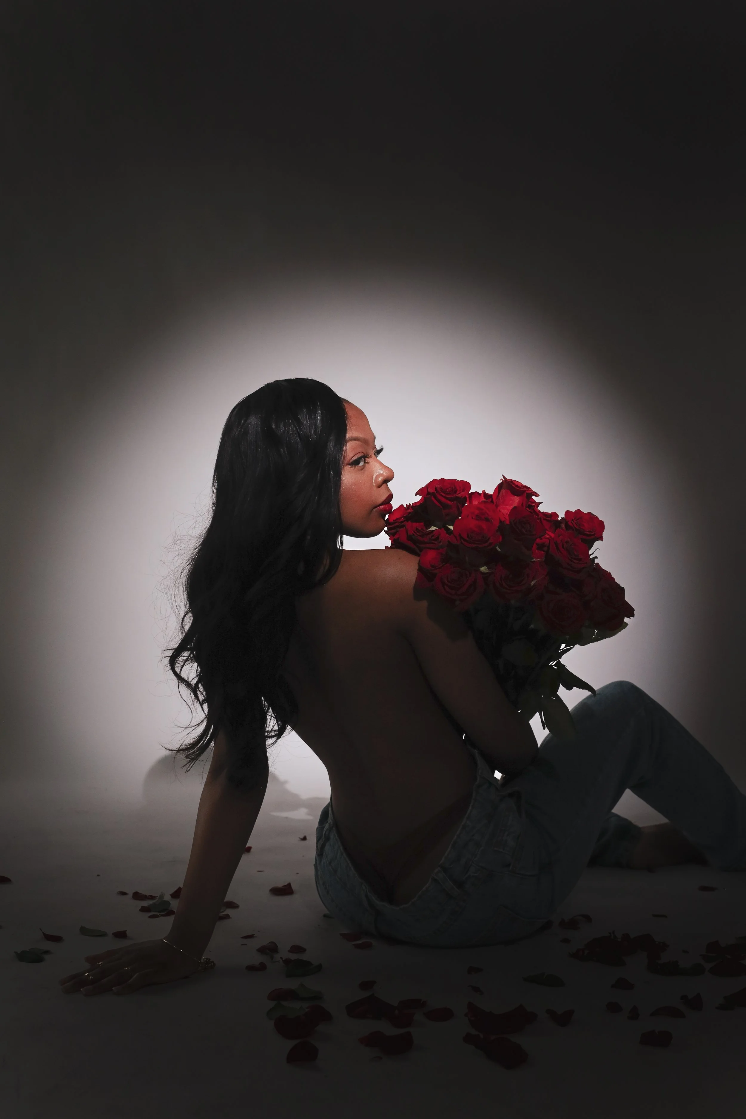 A woman with long black hair sitting on the floor, topless, holding a large bouquet of red roses, with scattered rose petals around her, against a gradient dark background.