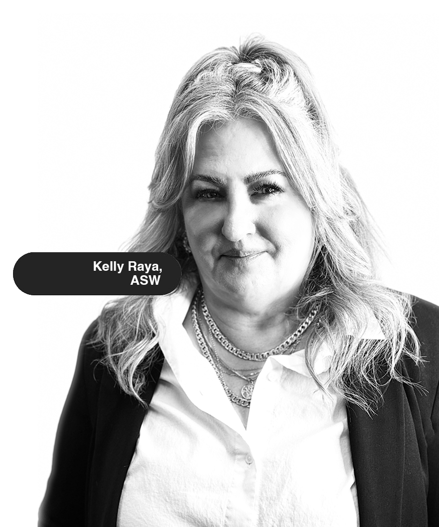 Black and white portrait of Kelly Raya, a woman with shoulder-length wavy hair, wearing a white collared shirt, layered necklaces, and a blazer, against a plain background. Victoria Community Counseling, LCSWC - Riverside, CA 