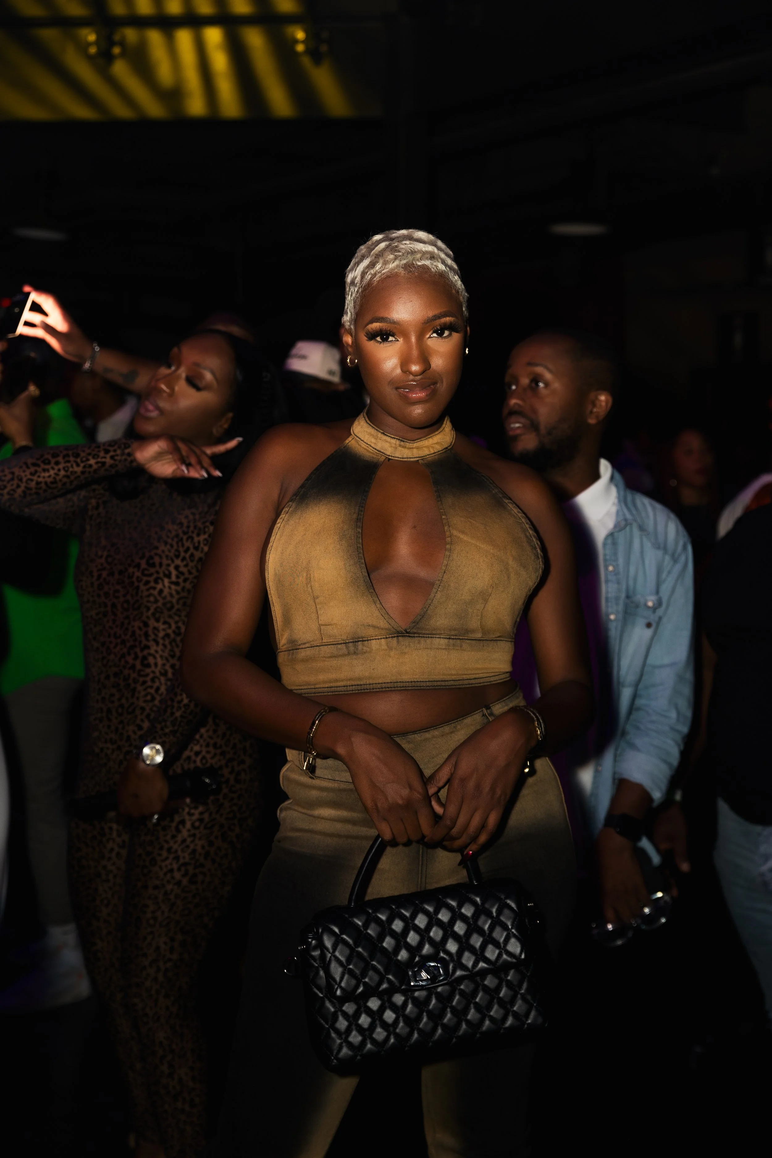 A woman with platinum blonde hair wearing a sleeveless, metallic gold top and matching pants holding a quilted black handbag at a dark event with other people in the background.
