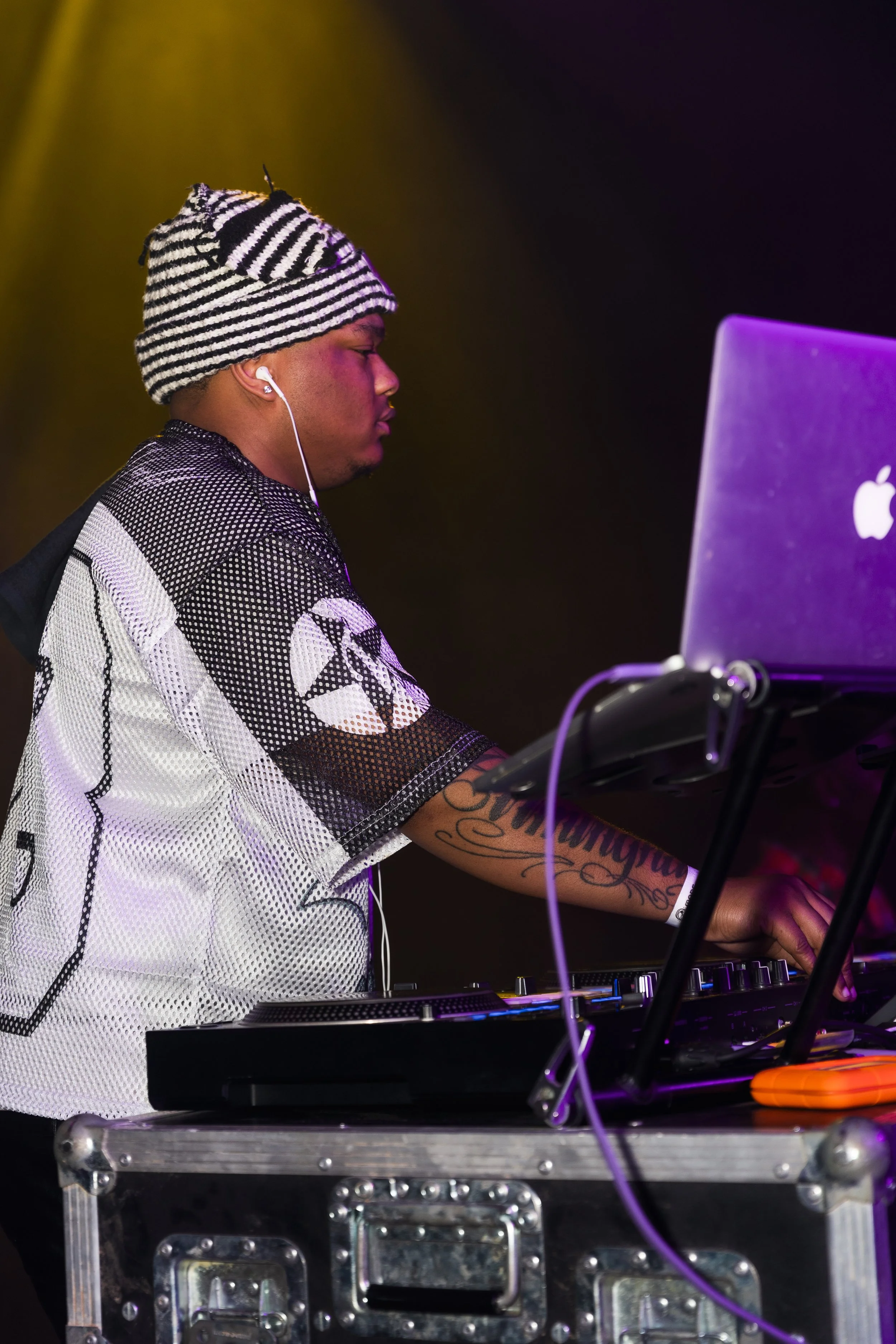 A DJ wearing a striped beanie and a black and white patterned shirt, using DJ equipment at a performance.