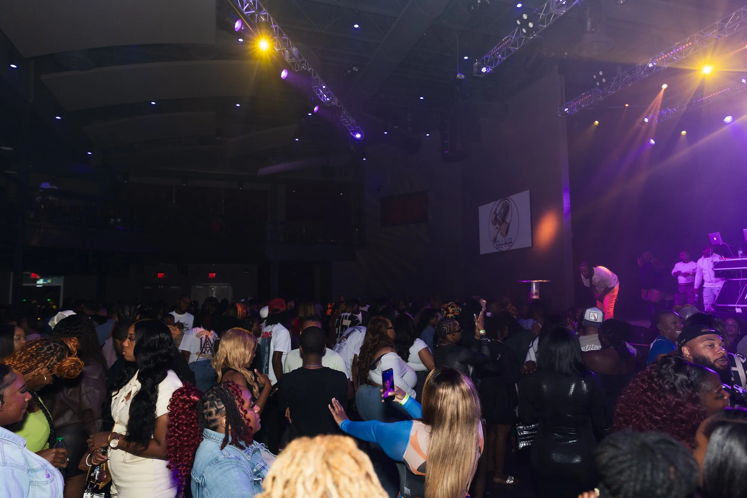 Crowd of people at a concert or nightclub, with a stage on the right featuring performers and DJ equipment, blue and purple lighting, and a large logo on the wall.
