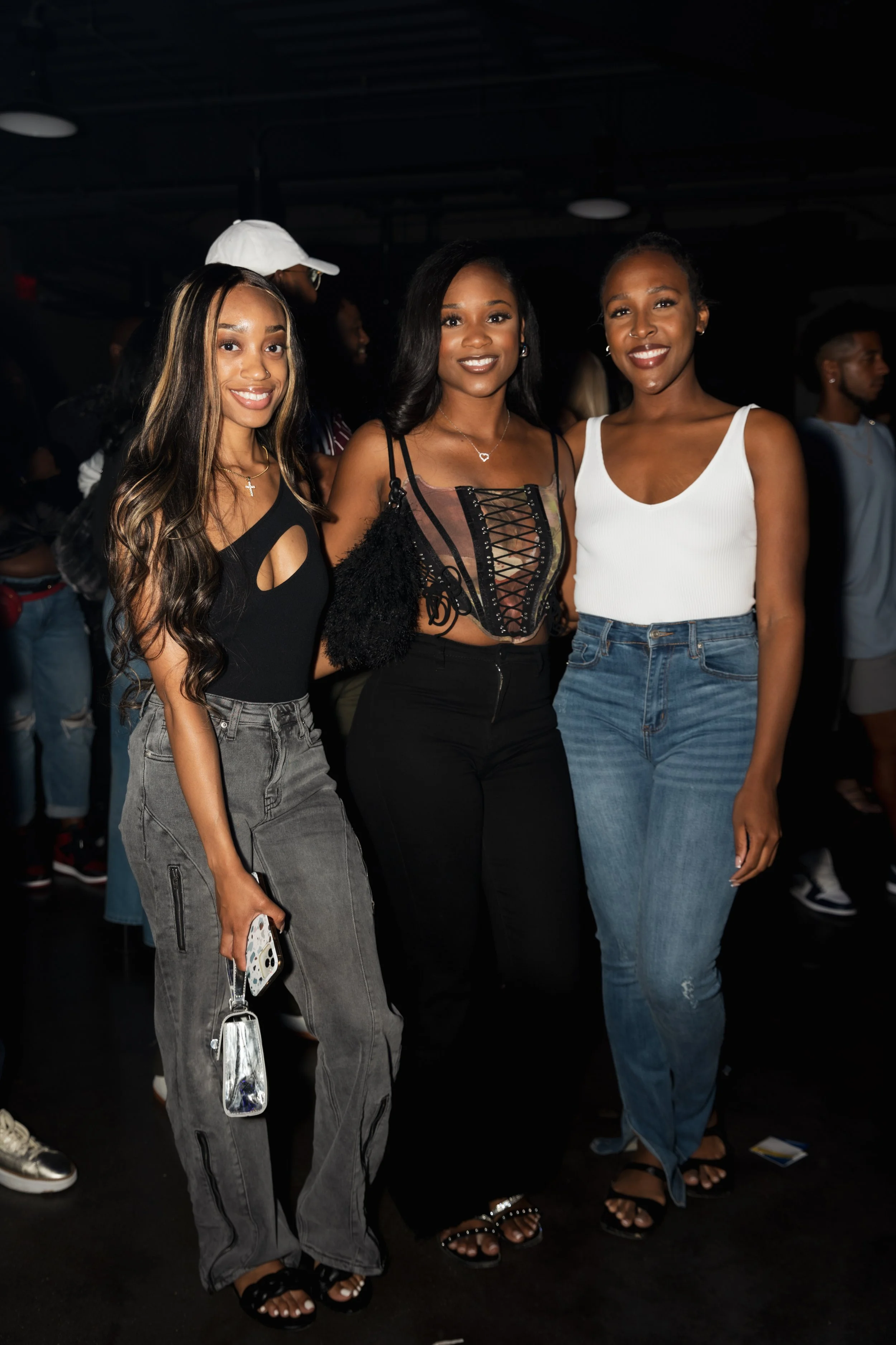 Three women standing together at an event, smiling at the camera, with a dark background and other people in the distance.