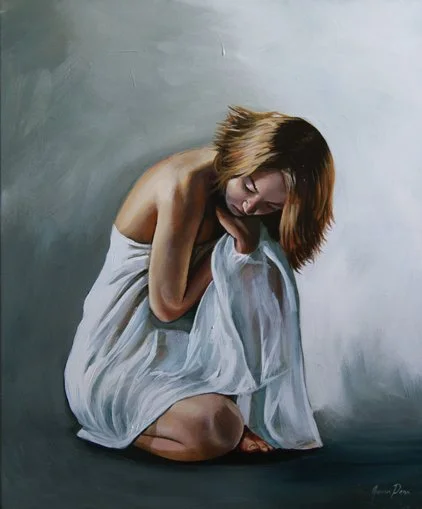 A woman with shoulder-length hair sitting on the floor, hugging her knees, wearing a white dress, and looking down in a contemplative pose.