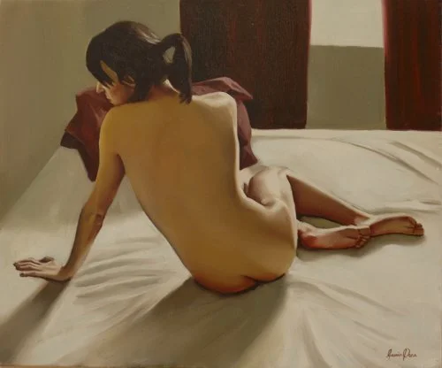 A woman with dark hair sitting on a bed, facing away, with pillows behind her head and a wall in the background.