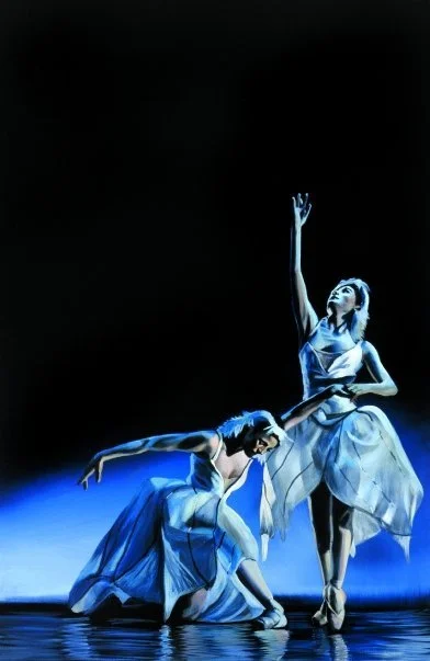 Two ballet dancers performing on stage, one is kneeling and bending forward, the other is standing with one arm raised, illuminated by blue stage lighting.