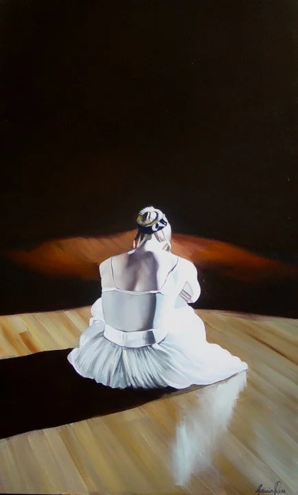 A person with dark hair sitting on a wooden floor, facing away, wearing a white dress and head covering, in a dimly lit room with dark background.