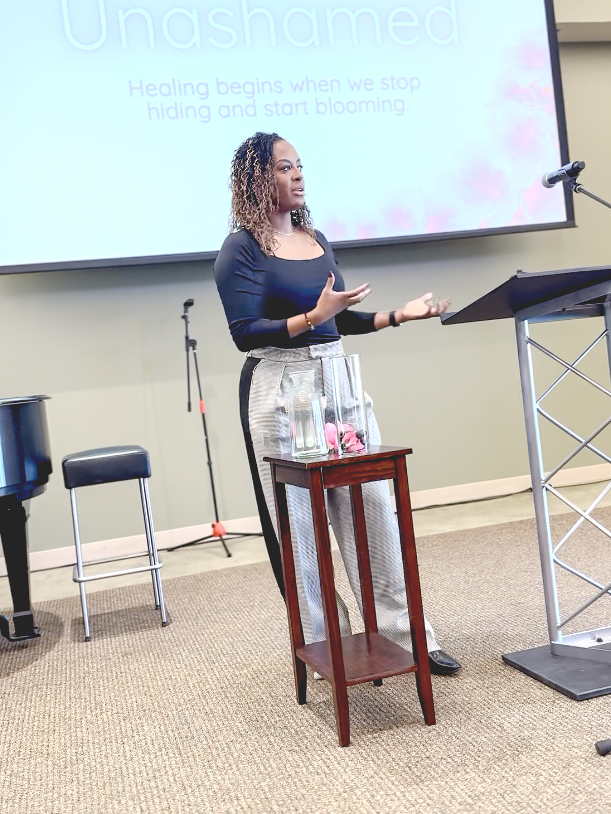 A woman giving a presentation in a conference room, standing near a podium with a glass water pitcher and glasses, with a large screen behind her displaying the word 'Unashamed' and the phrase 'Healing begins when we stop hiding and start blooming.'