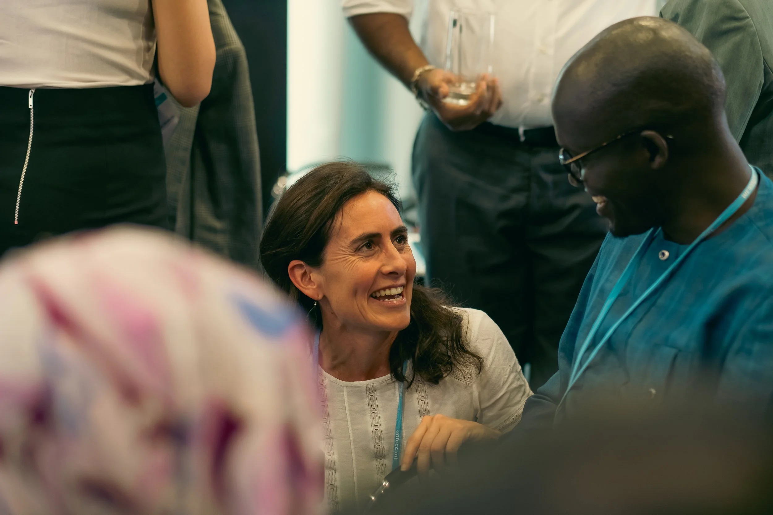 A woman with dark hair smiling and looking at a man in glasses, who is also smiling, during a conversation in a group setting.
