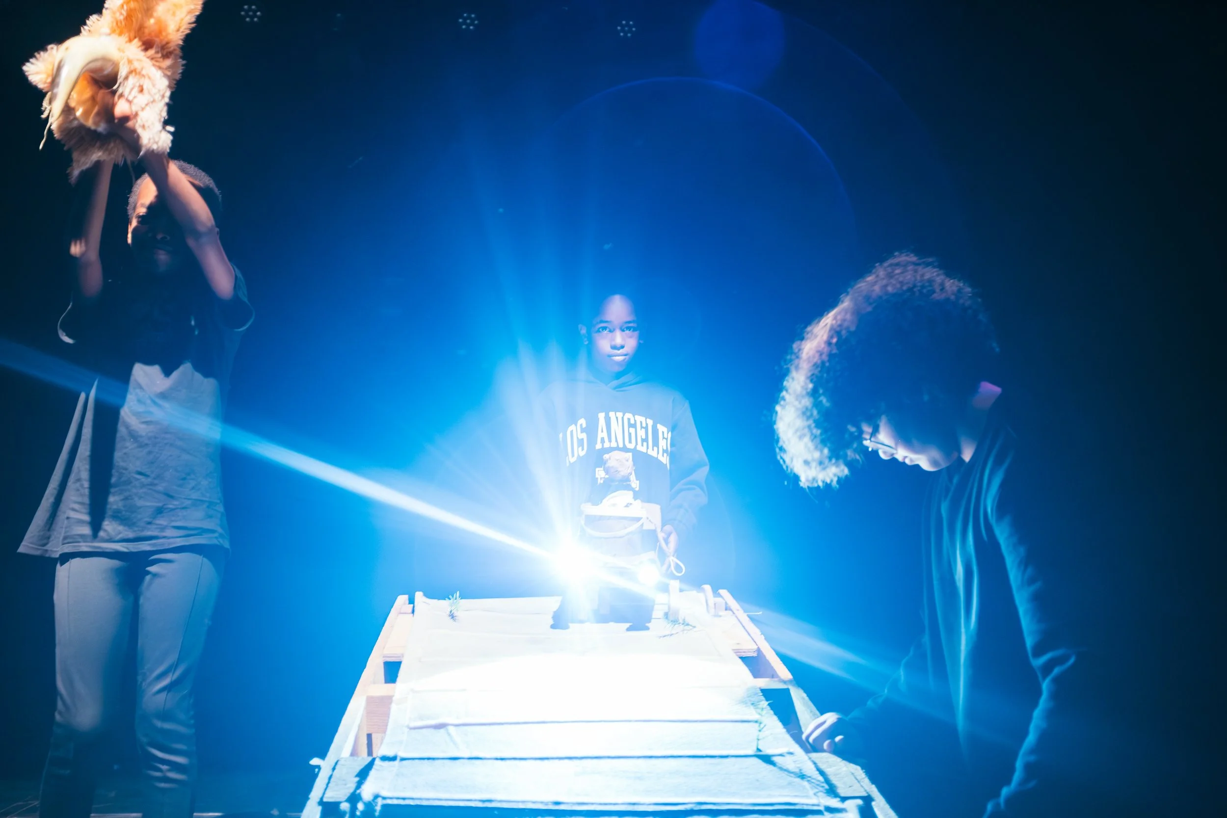 Three children stand around a table with a bright light source. One child, on the left, holds a fluffy object above their head. The child in the middle, wearing a Los Angeles hoodie, is illuminated by the light, and the child on the right, with curly