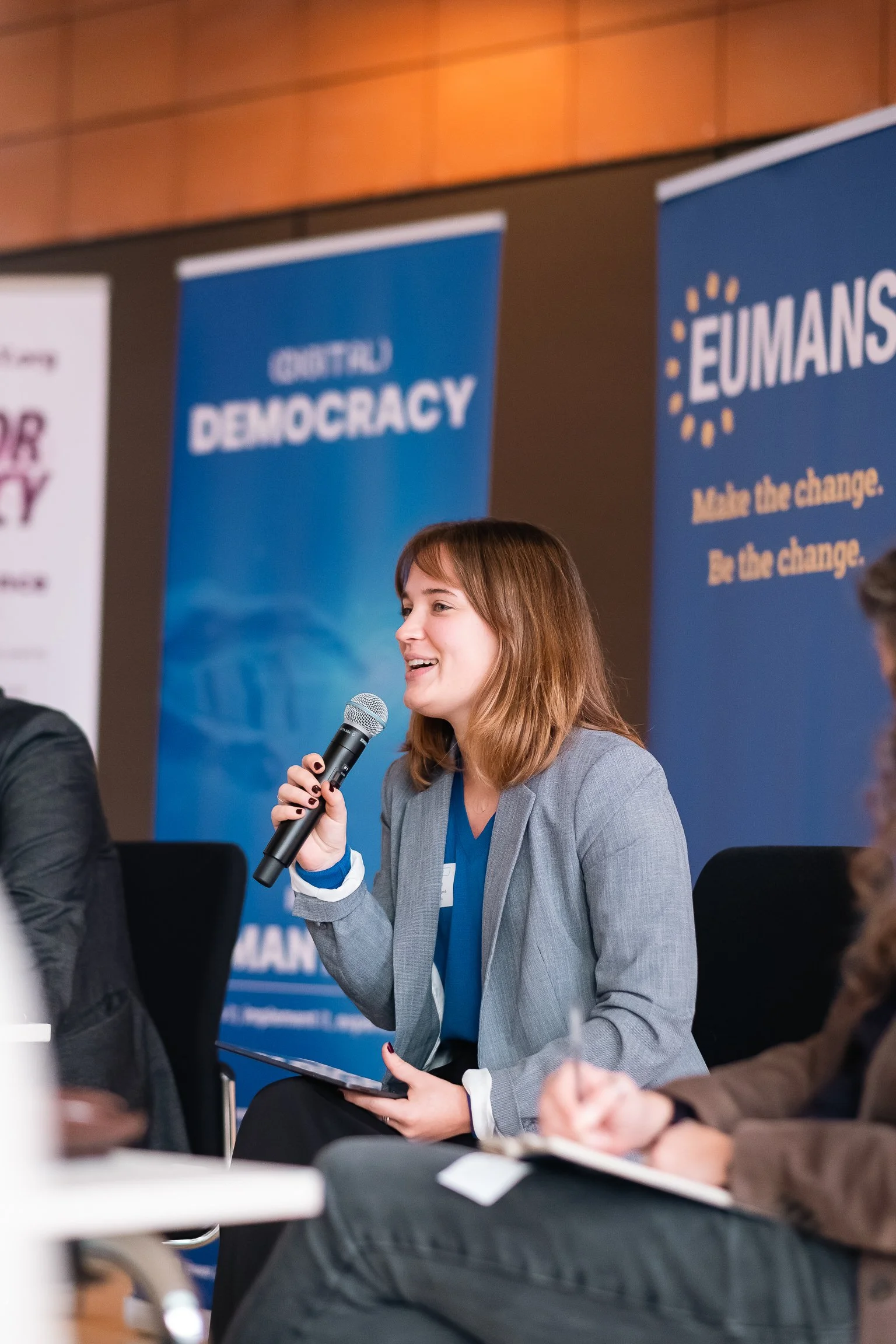 Young woman with shoulder-length brown hair speaking into a microphone at a conference. She is dressed in a gray blazer and blue top, seated with an audience in front of banners behind her that read "CENTER FOR DEMOCRACY" and "EUMANS."