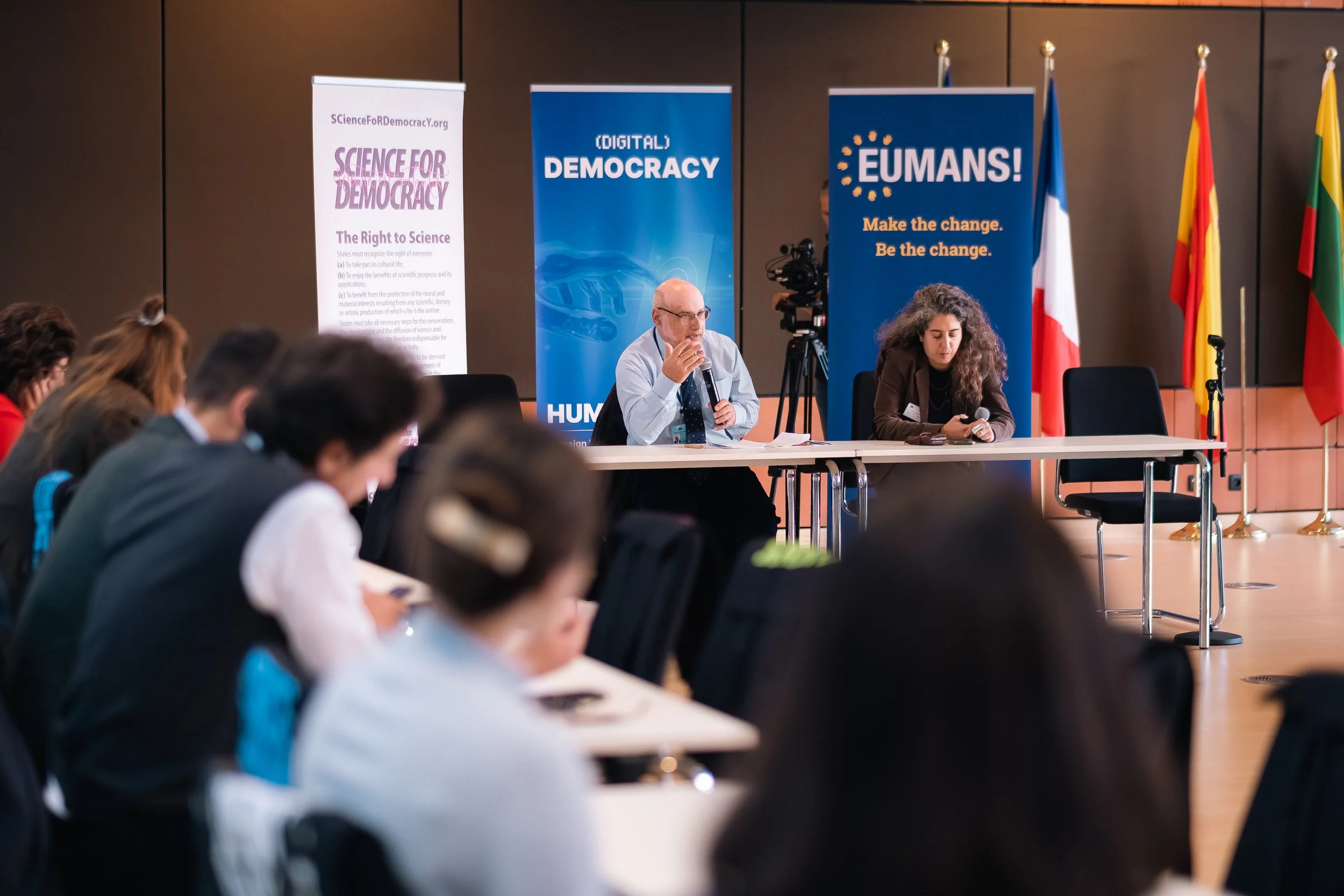 A conference room with a panel of two speakers at a table in front of an audience. Behind them, banners with the words 'Science for Democracy,' 'Digital Democracy,' and 'EUMANS!' are visible, along with flags.