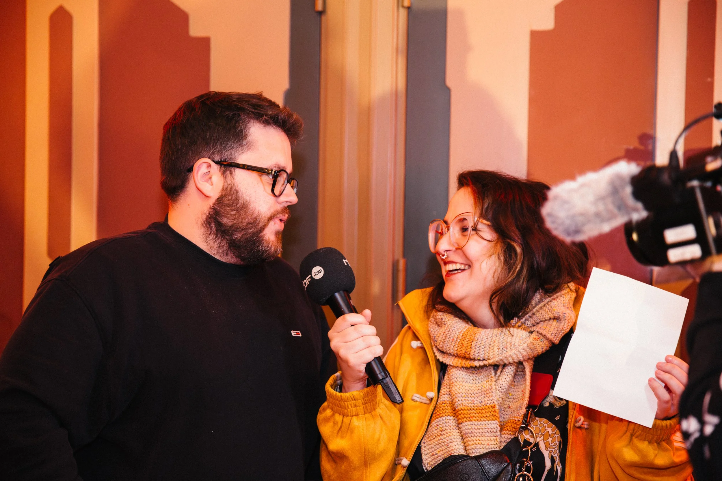 Two people being interviewed, one man with glasses and a beard holding a microphone, and a woman with glasses and a scarf, smiling and holding a piece of paper, inside a room with wooden paneling.