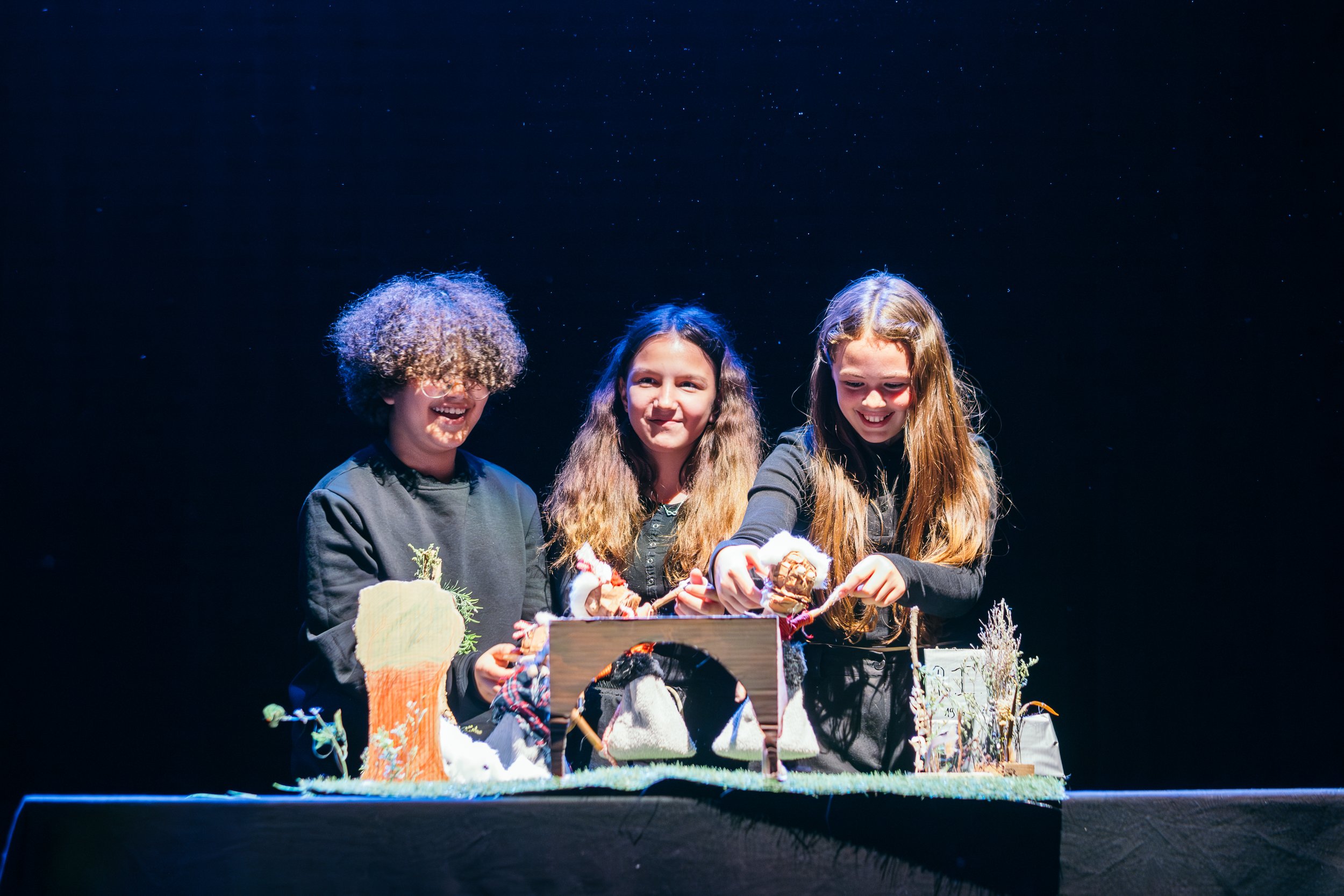 Three children with long and curly and straight hair play with a craft project on a table, with a dark background and spotlight.