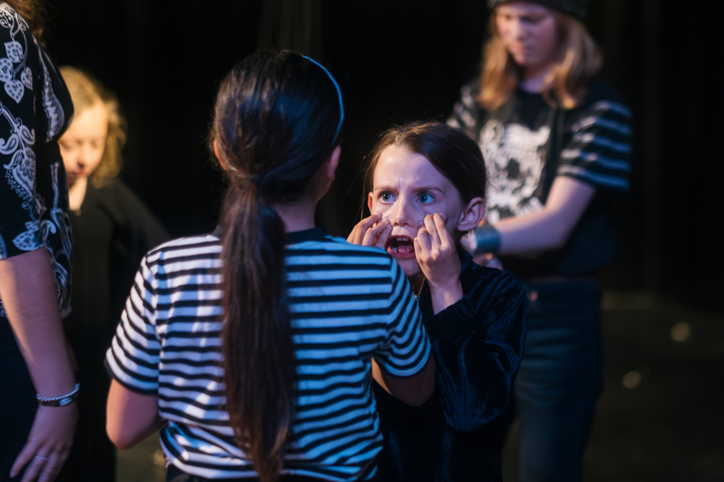 A girl with long dark hair and in a black outfit appears distressed, with her hands on her face, while another girl with long dark hair and a striped shirt holds her face and seems to be comforting her, in a dark setting with other girls in the backg