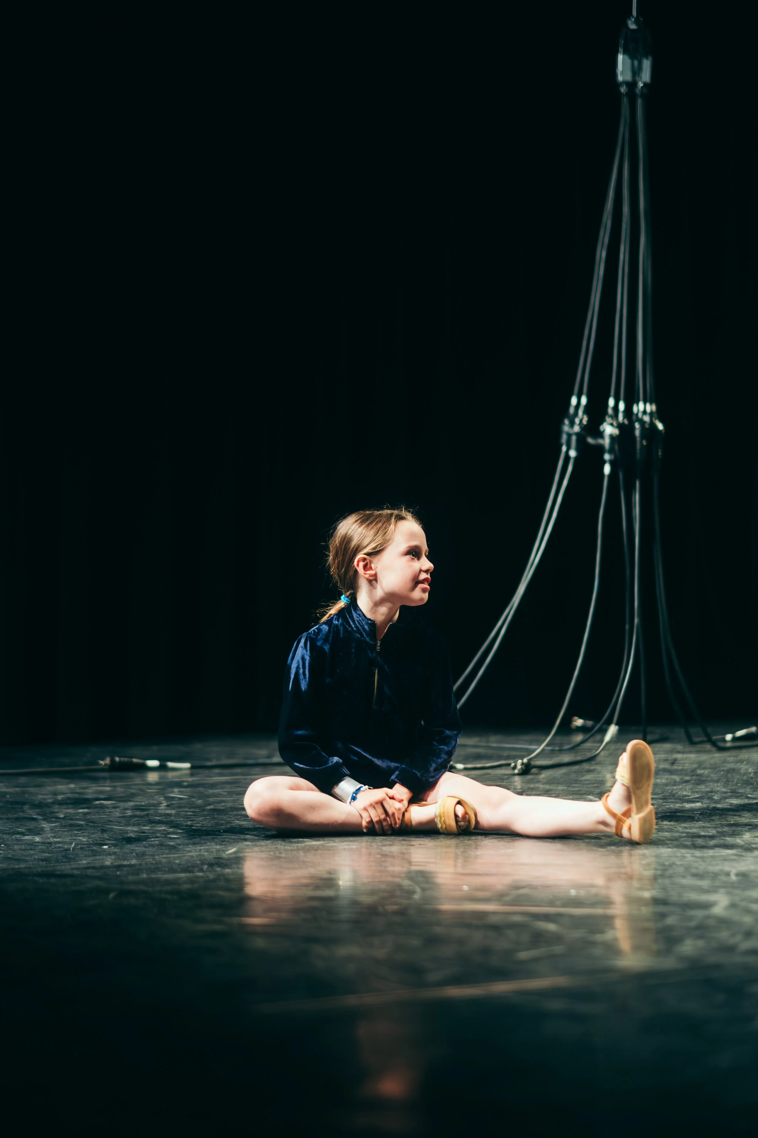 A young girl in a blue jacket sitting on a stage with black curtains, stretching her legs with her right hand resting on her ankle and her left hand holding her foot.