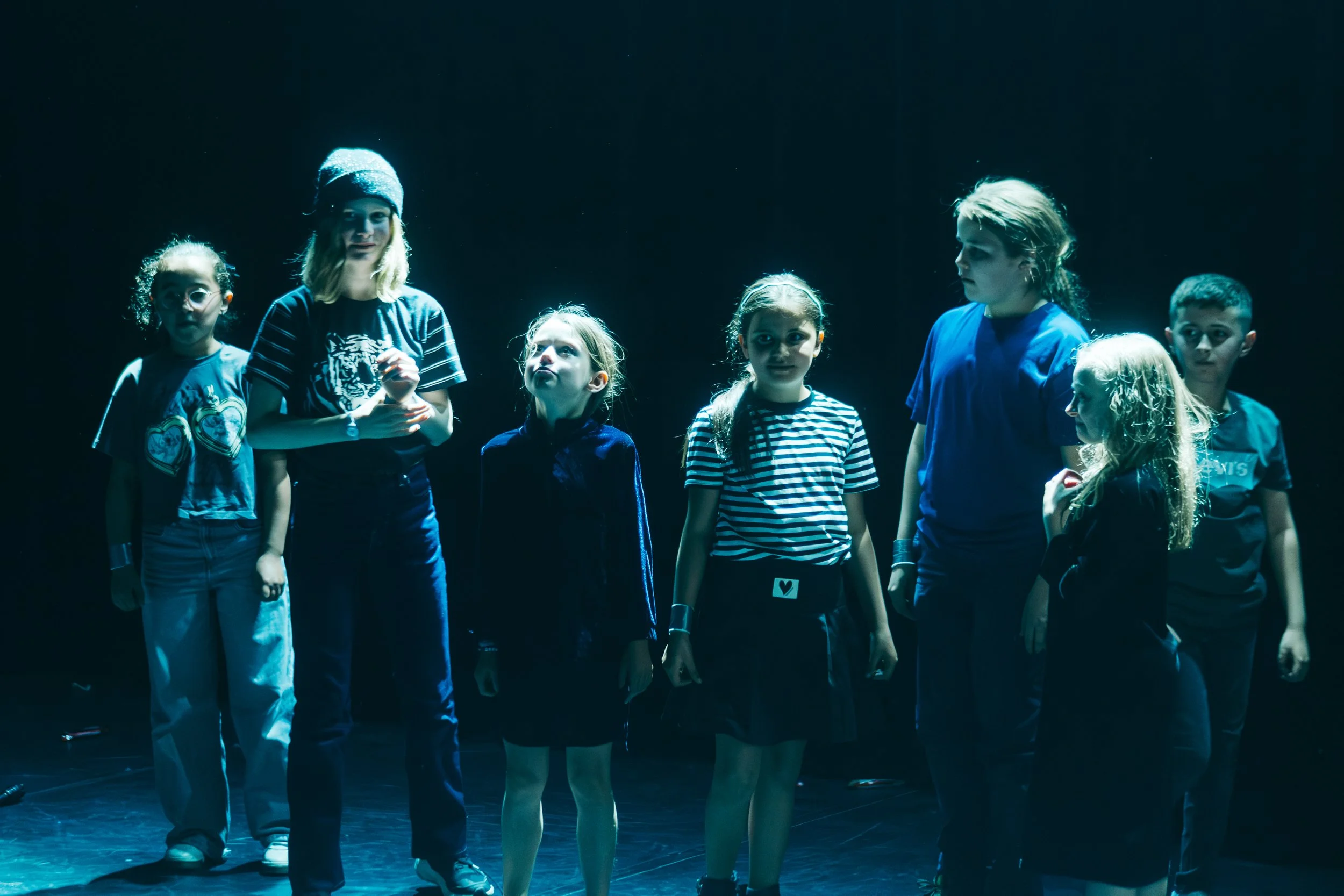 Children standing on stage under a spotlight, dressed in casual clothes, at a performance or rehearsal.