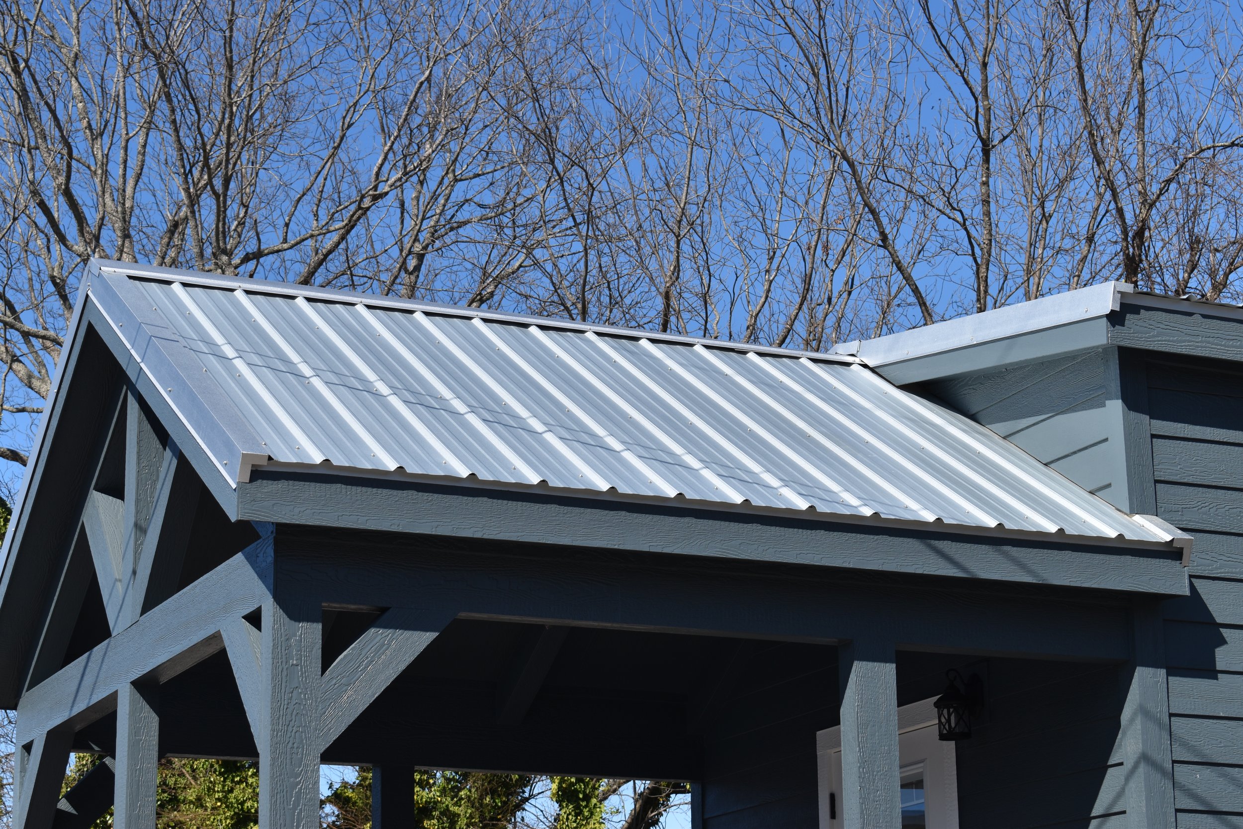 A close-up of a small metal roof with a house exterior featuring blue-gray siding and a black lantern-style light fixture on a clear day with bare tree branches and a blue sky in the background.