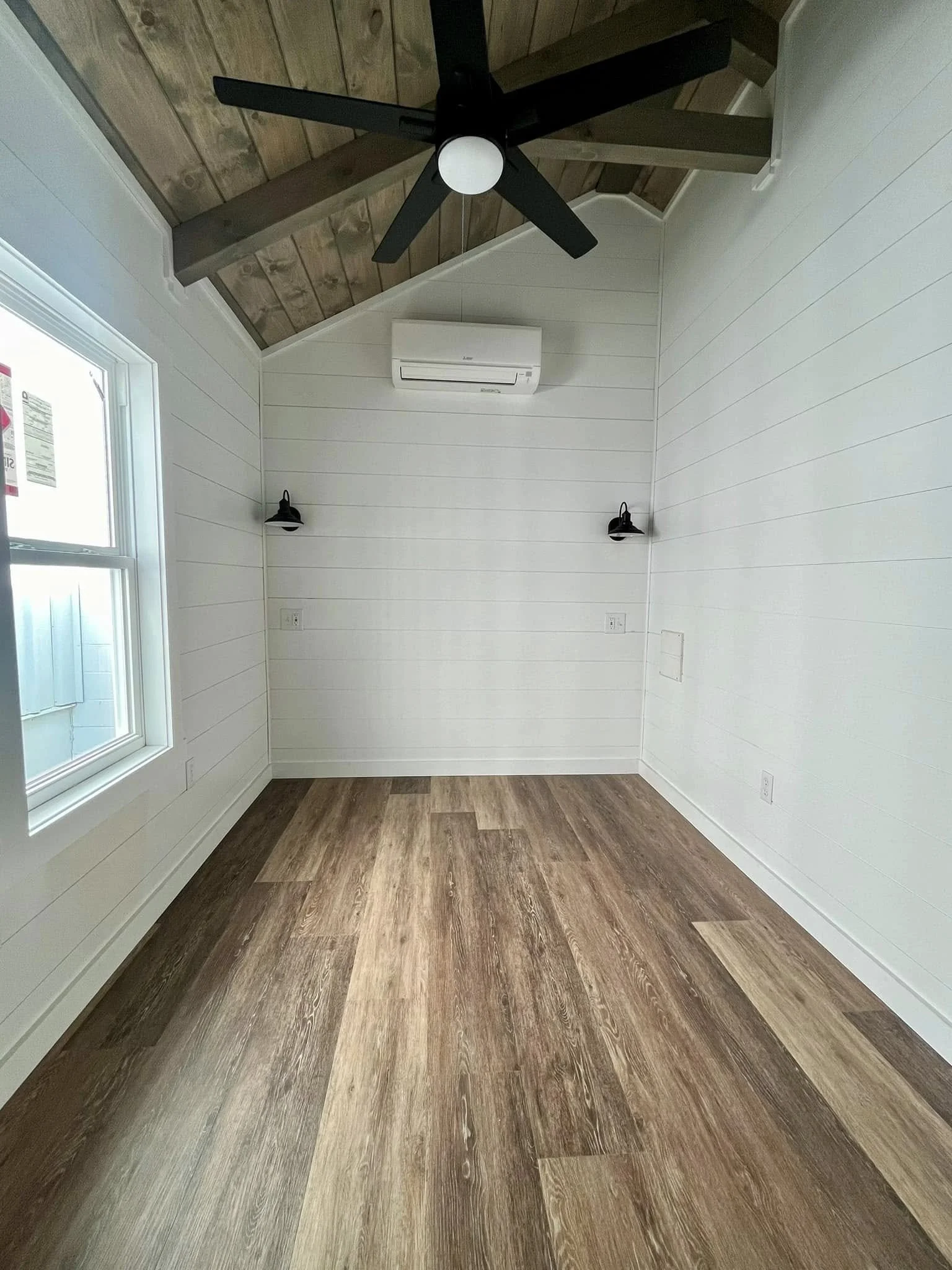 Empty room with white shiplap walls, hardwood floors, a window on the left, two black wall-mounted lamps, a white air conditioning unit, a ceiling fan with black blades, and a sloped wooden ceiling.