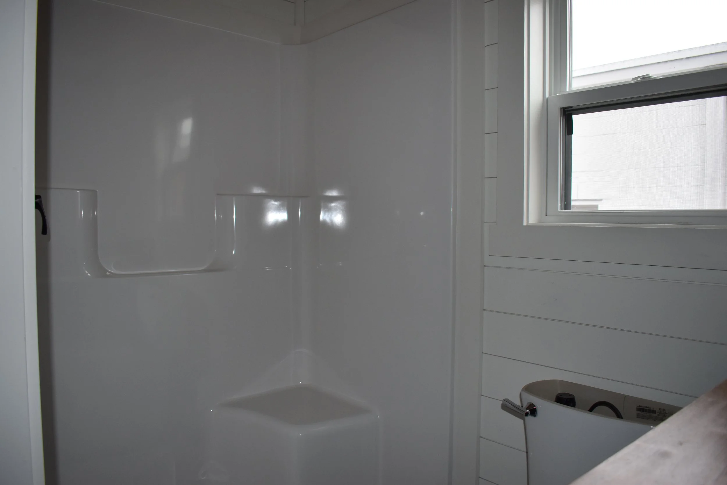 Empty white shower stall with built-in soap dish and a window in a white-paneled bathroom.