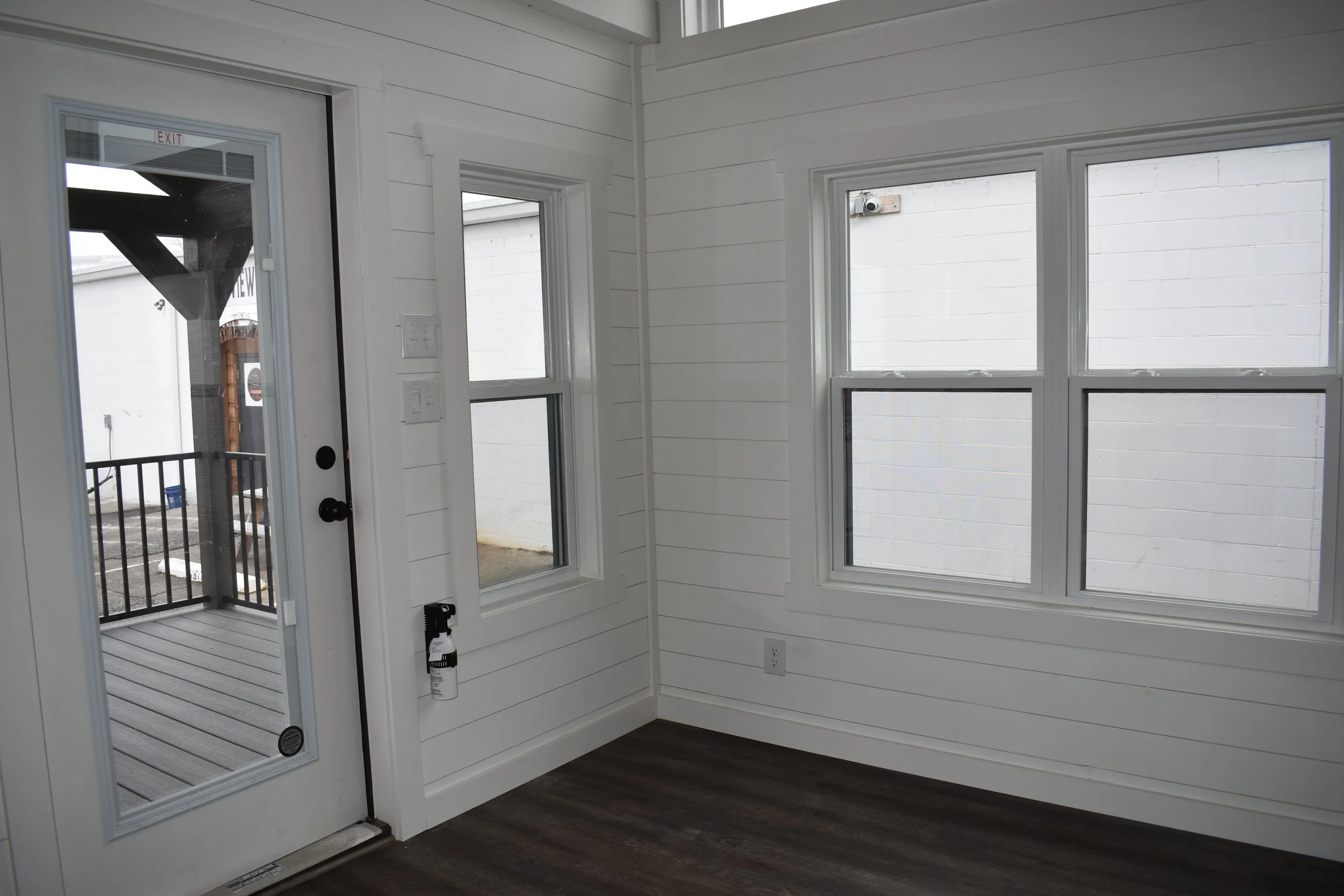Interior of a newly built room with white walls, three windows, a door with a glass panel opening to an outdoor deck, and dark wood flooring.