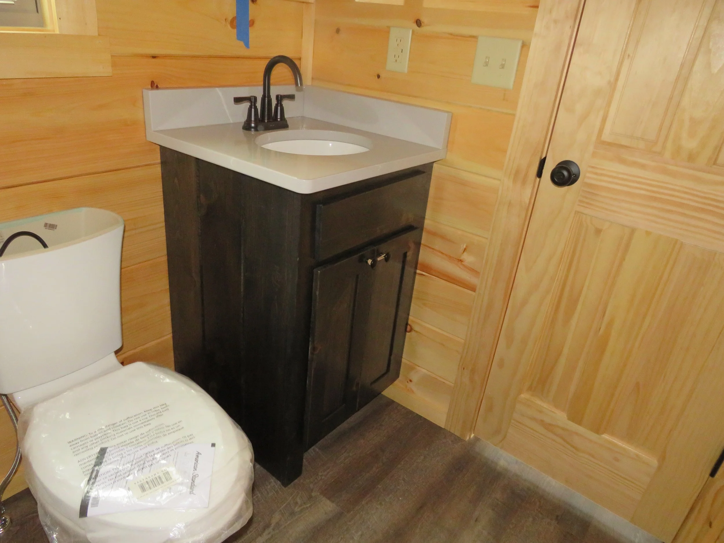 Bathroom with wood-paneled walls, a small black vanity with a white sink and black faucet, a white toilet, and a wooden door.