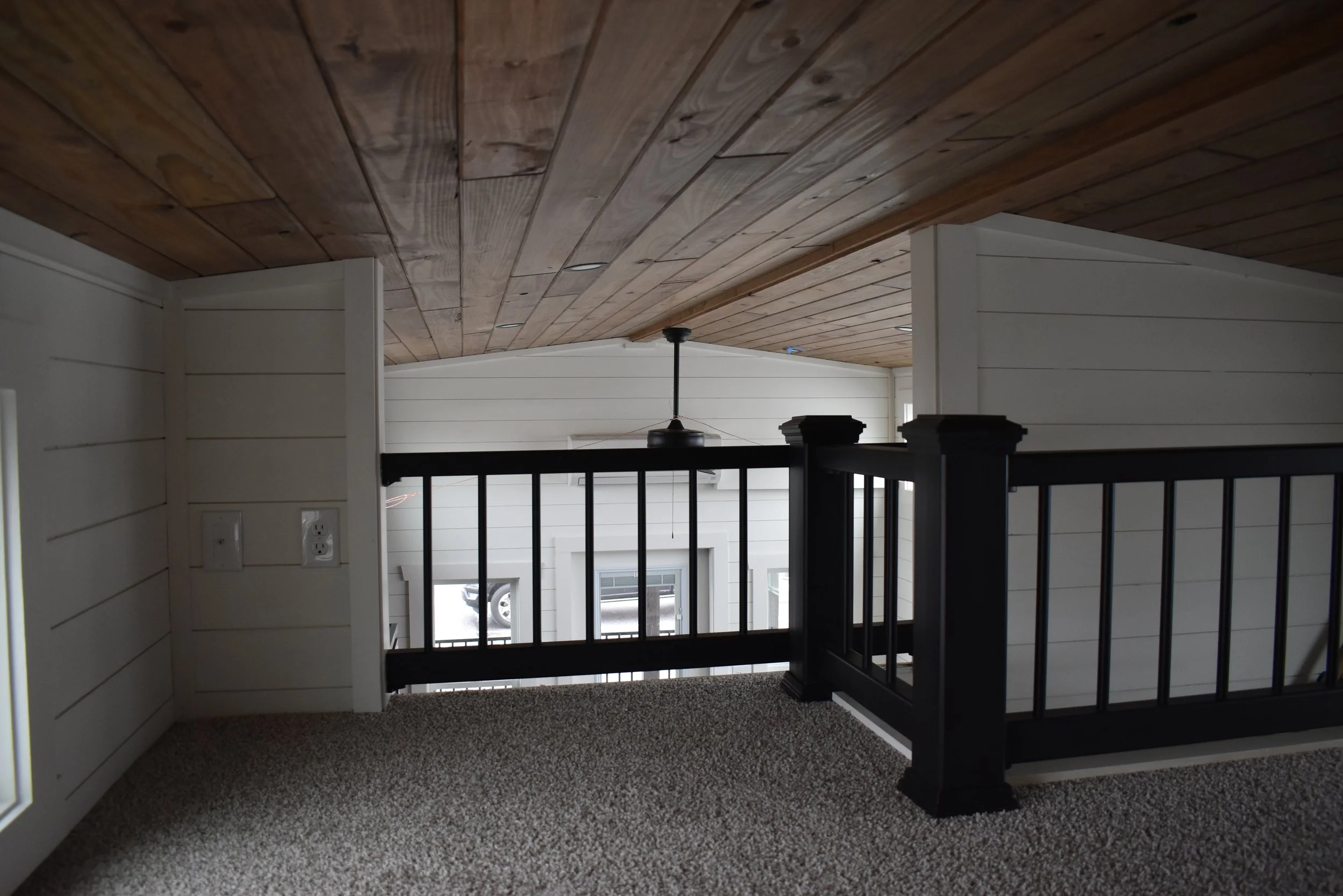 Interior view of a second floor or balcony with black wooden railing, beige carpeted floor, white paneled walls, and a wooden ceiling, overlooking a lower level with large windows and driveway.