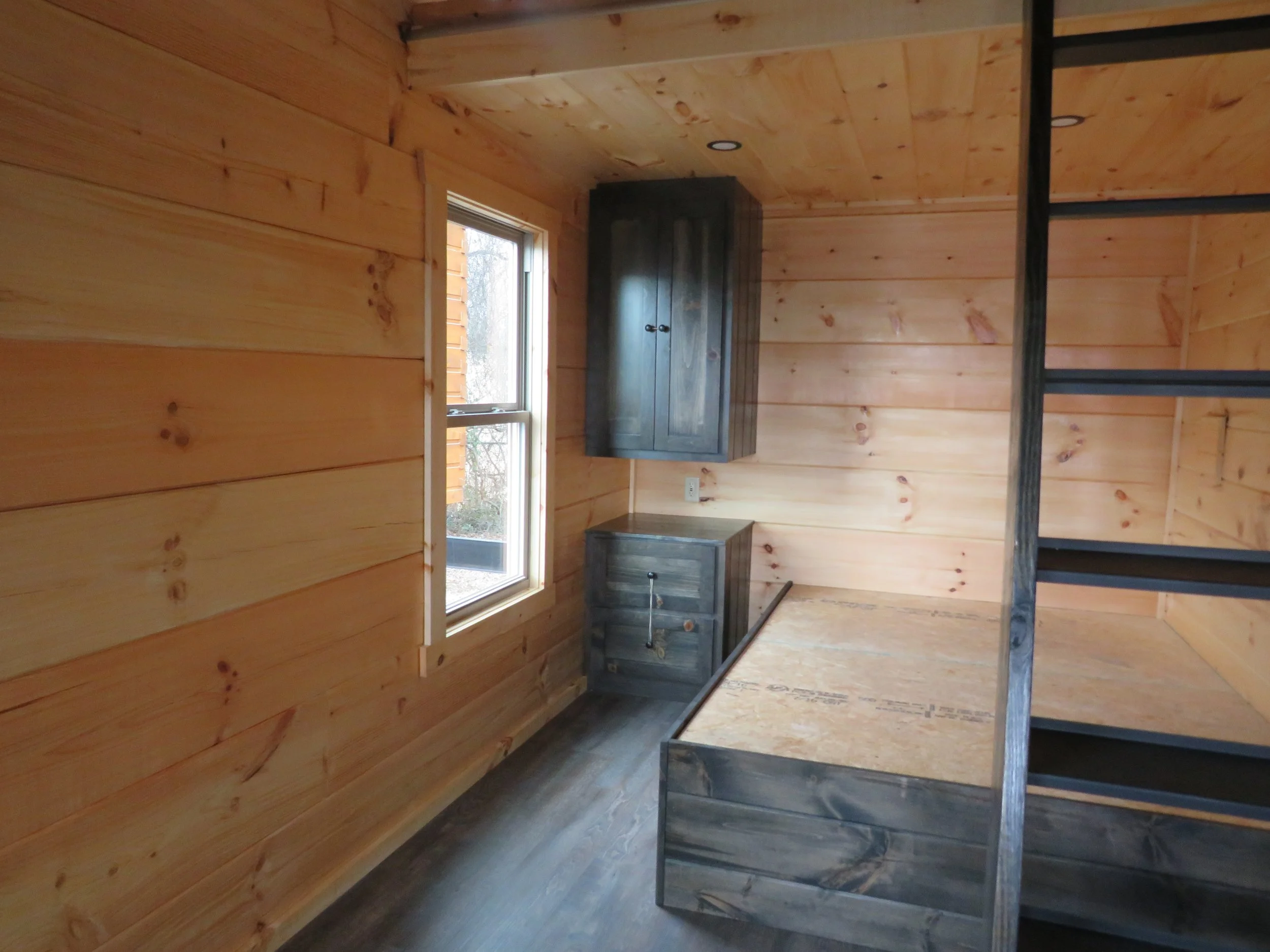 Room with natural wood paneling walls, a window, a dark gray cabinet and dresser, and partially assembled bunk bed frame.