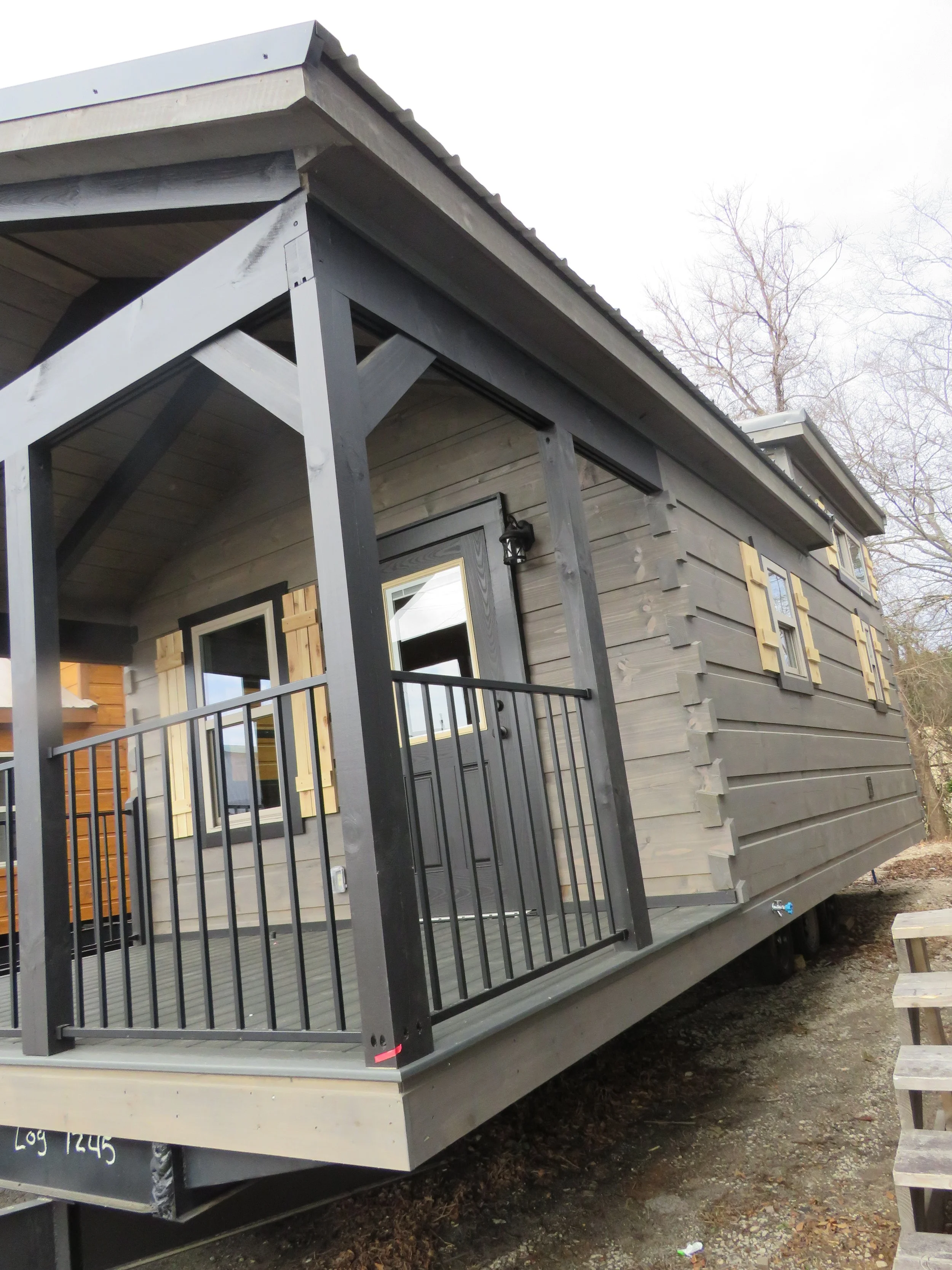 A small modern house with a covered porch, black railing, gray siding, and multiple windows with light-colored wooden shutters.