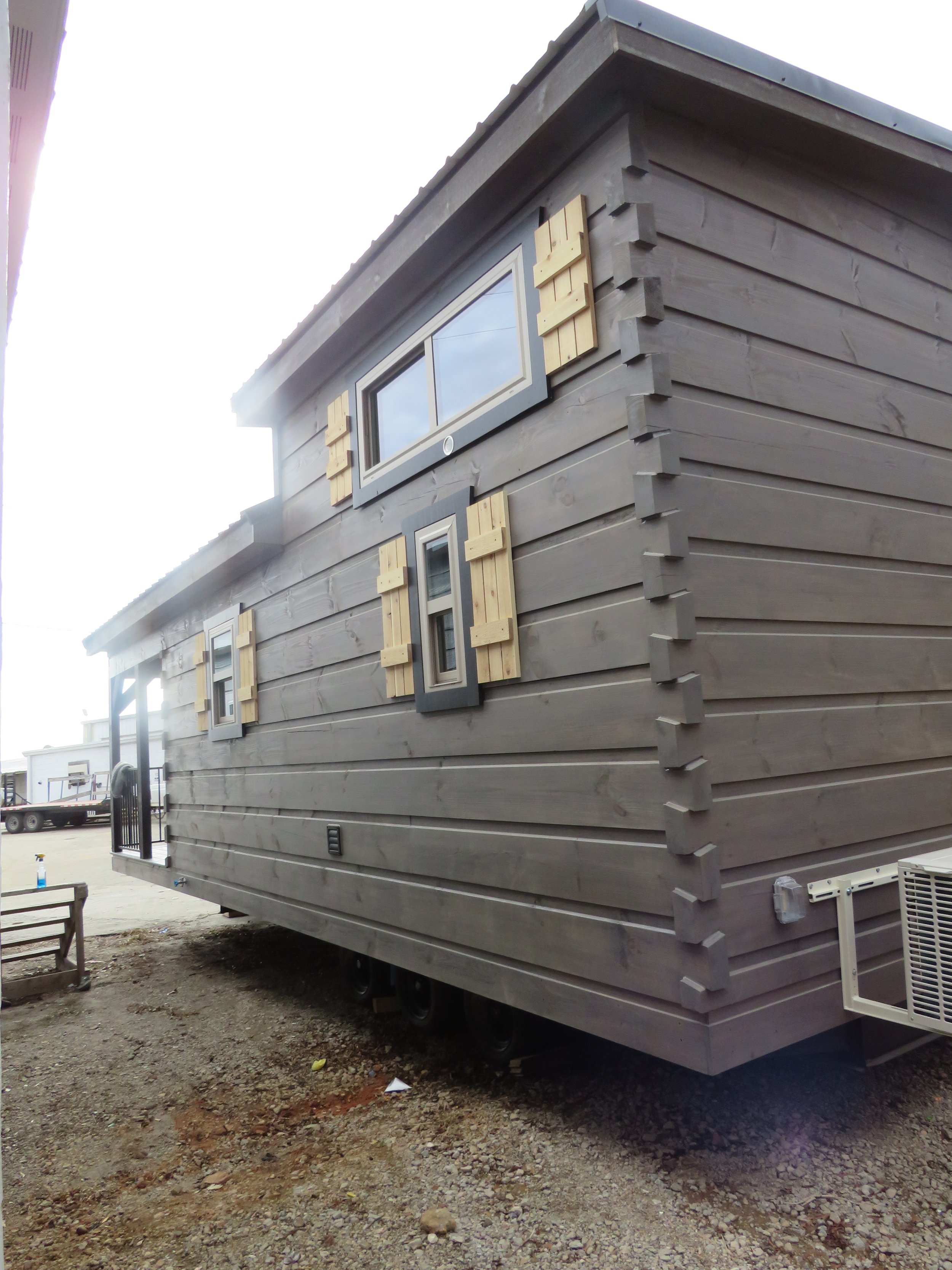 A tiny house on wheels with dark gray wooden siding and three small windows, each with light-colored wooden shutters. The house is elevated above the ground on trailer wheels.