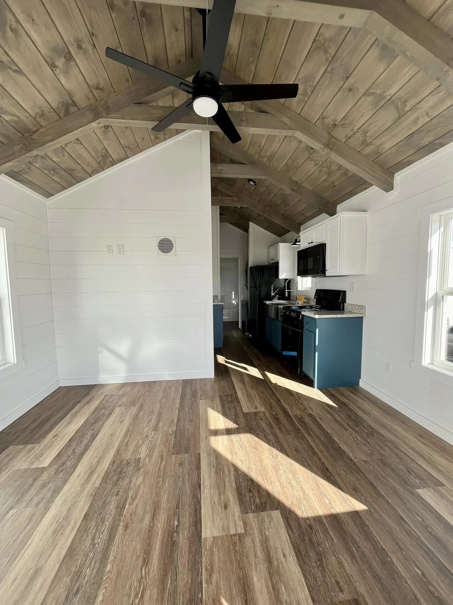 Empty room with wood flooring, white walls, a wooden vaulted ceiling with a black ceiling fan, and a kitchen area with black appliances and teal cabinets.