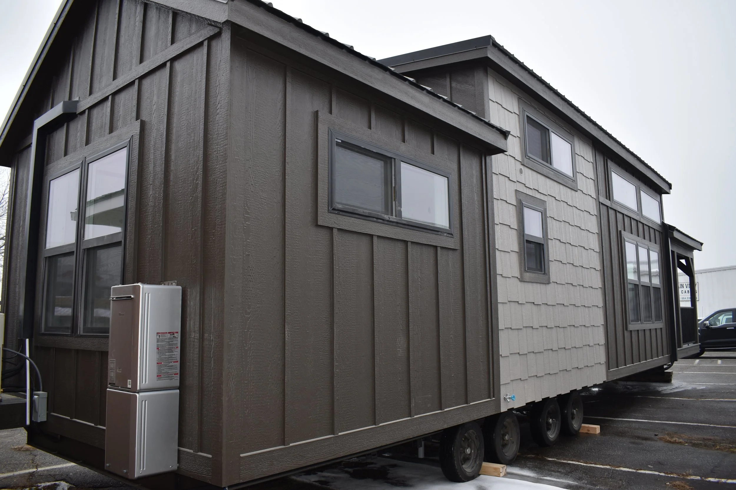 A tiny house on wheels with dark brown and light gray exterior siding, multiple windows, and a propane tank attached to the side, parked in a parking lot.