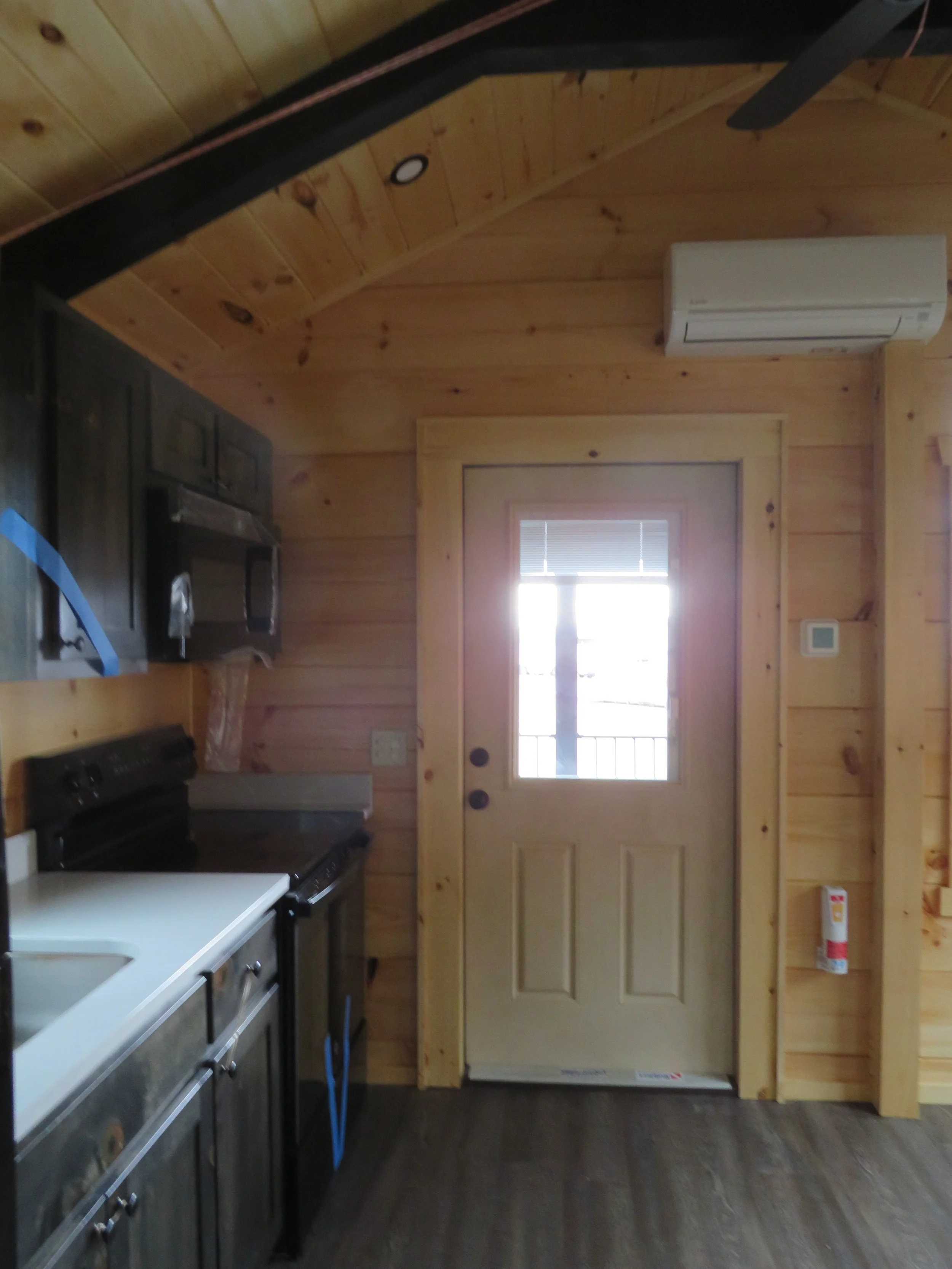 Interior view of a wood-paneled kitchen with a door to outside, black cabinets, microwave, stove, and laminate flooring.