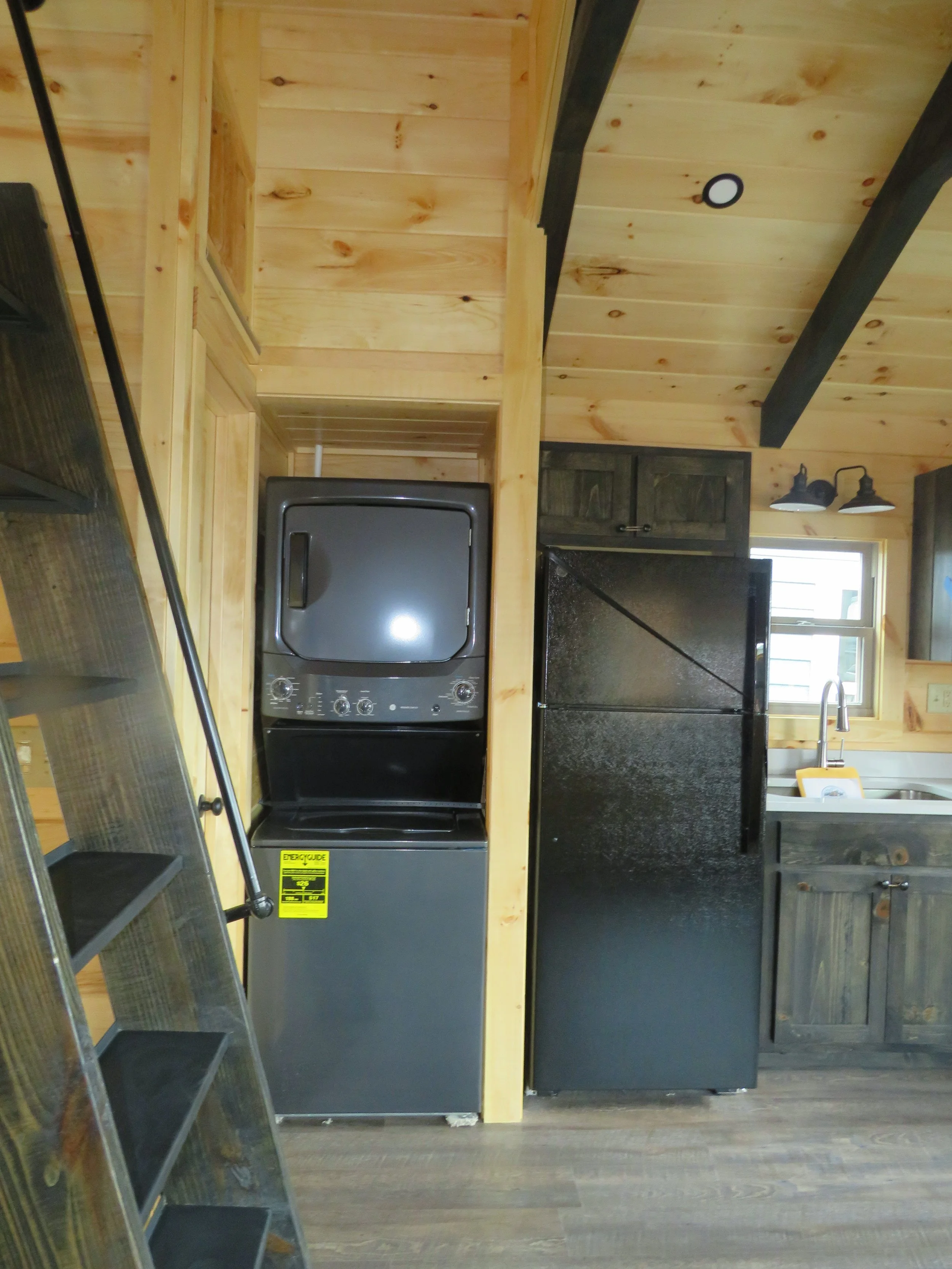 A small laundry and kitchen area with wooden walls and ceiling. On the left, there is a ladder. In the center, there is a stacked washer and dryer unit and a black refrigerator. On the right, there is a kitchen sink under a window with a yellow book 