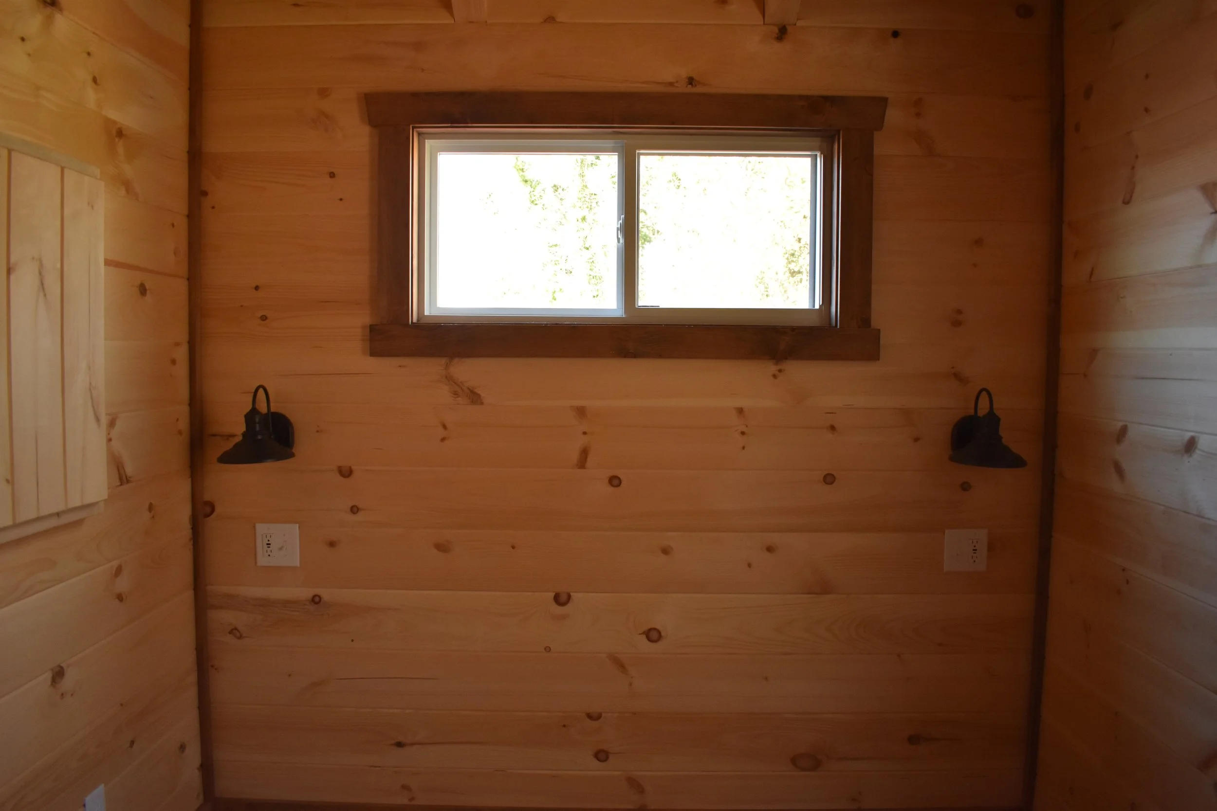 A small room with wooden walls, a rectangular window, and black wall-mounted lamps on either side of the window.