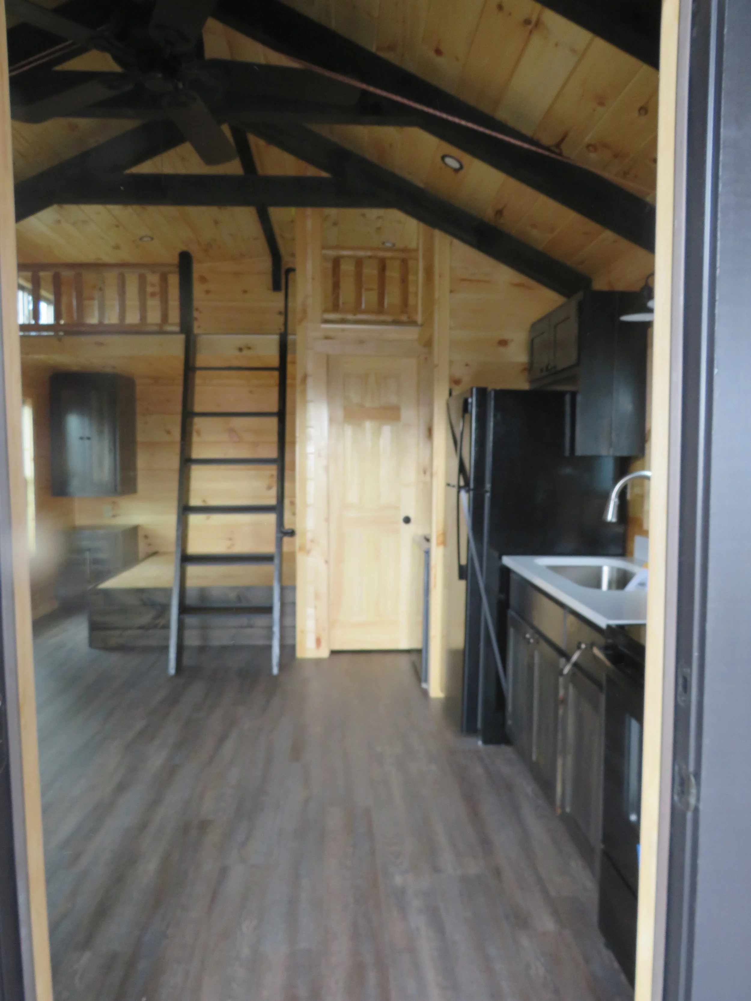 Interior of a small wooden cabin with a loft area accessed by a ladder, kitchen with black appliances, stainless steel sink, and wooden walls and ceiling.
