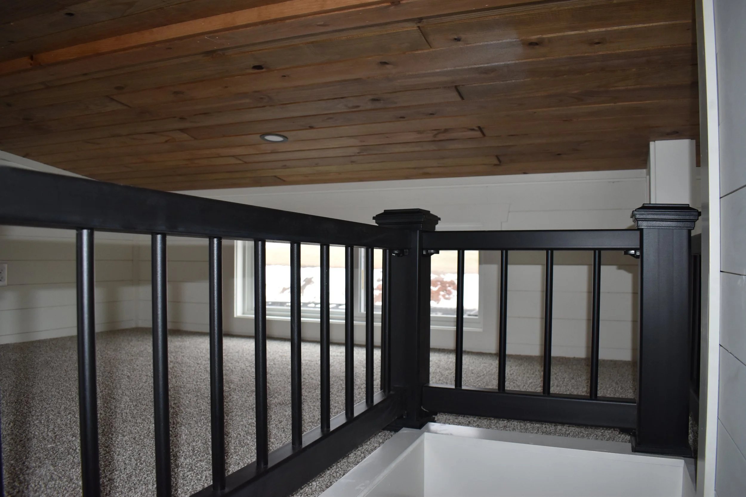 Interior view of a black wooden staircase railing, carpeted floor, and ceiling with wooden planks, in a room with white walls and a window showing a snowy landscape outside.