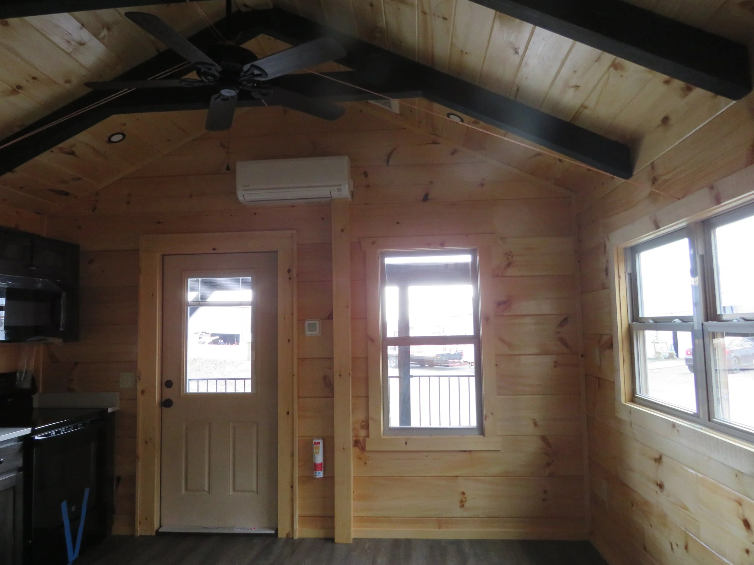 Interior of a small wood-paneled room with a ceiling fan, an air conditioning unit, a door with a window, and a window on the right wall, showing parked cars outside.