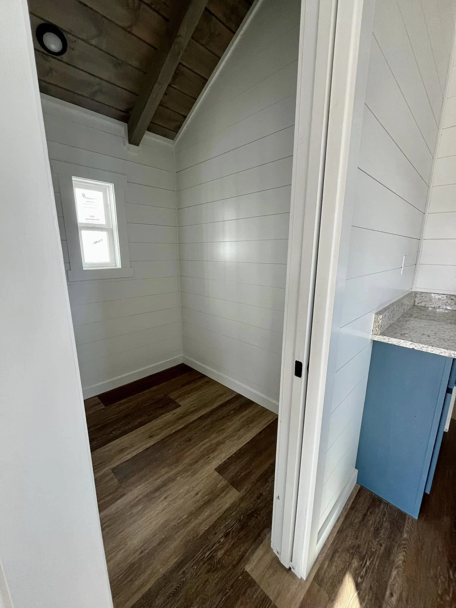 A small room with white paneled walls, wood flooring, a tiny window, and a sloped ceiling with wooden beams.