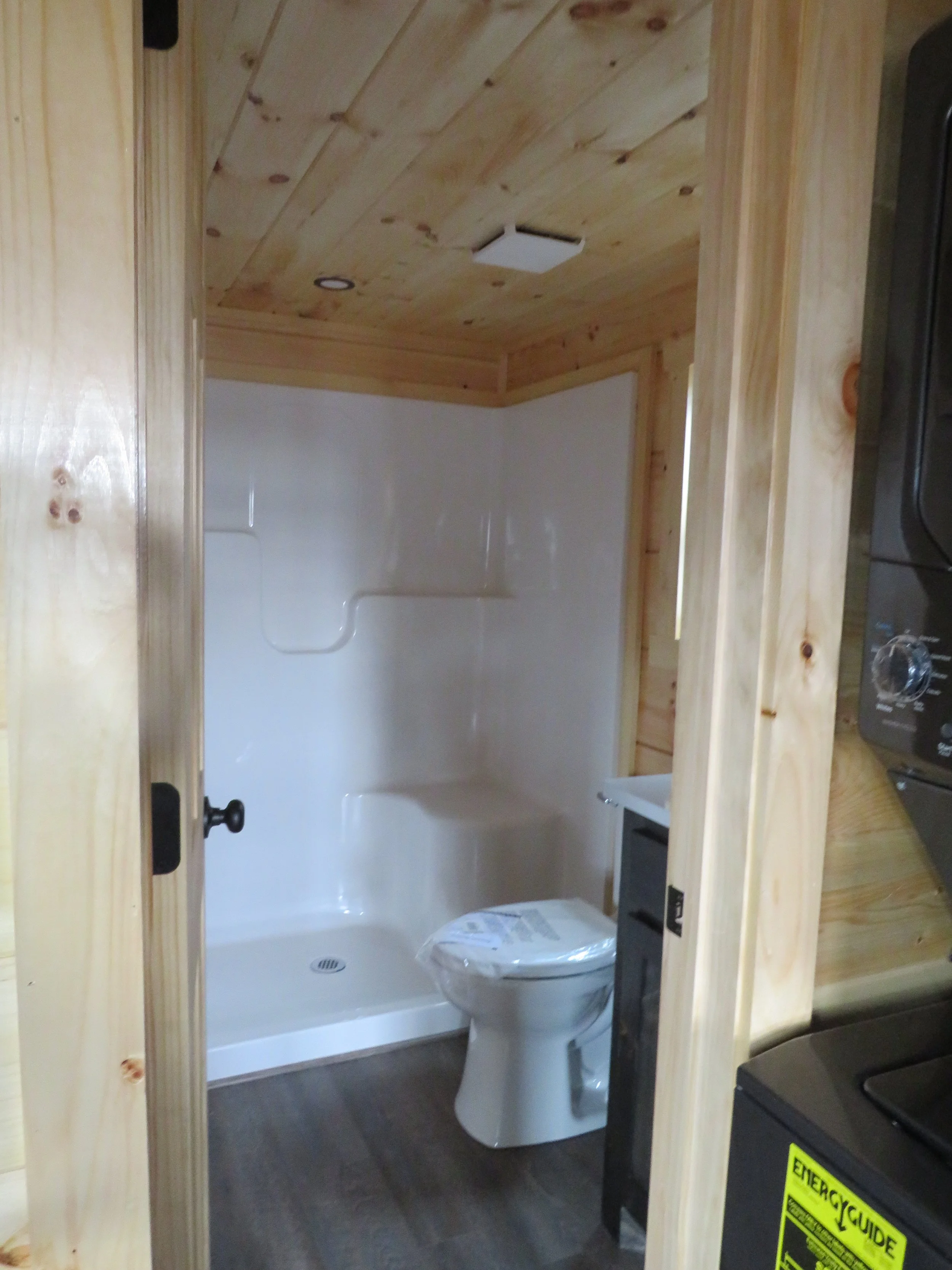 A small bathroom with a white shower and toilet, framed by unfinished wooden walls and ceiling. There are built-in shelves in the shower and a small black cabinet next to the toilet.