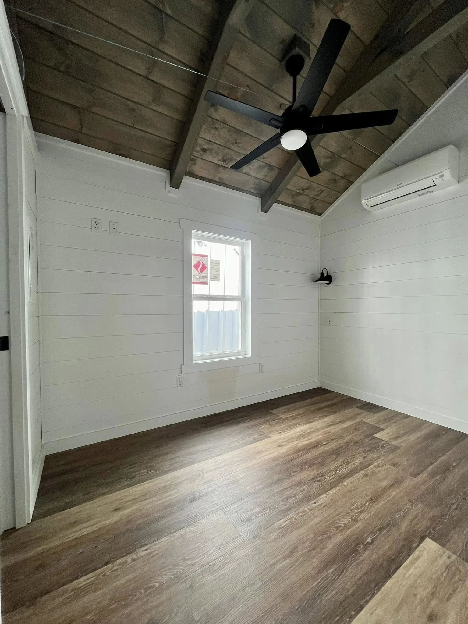 Empty room with wooden flooring, white shiplap walls, a small window, a black ceiling fan, a wall-mounted light, and an air conditioning unit.