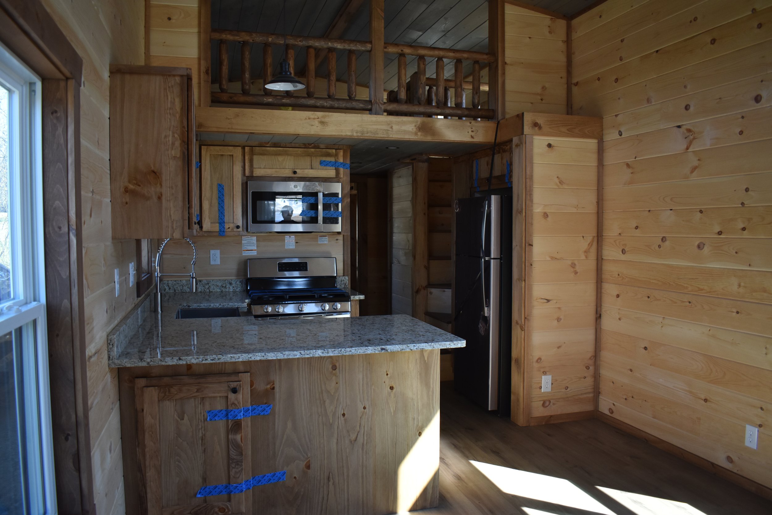 Interior of a wooden cabin kitchen with new appliances, granite countertop, and unfinished wood cabinets and walls; a loft area above with a railing.