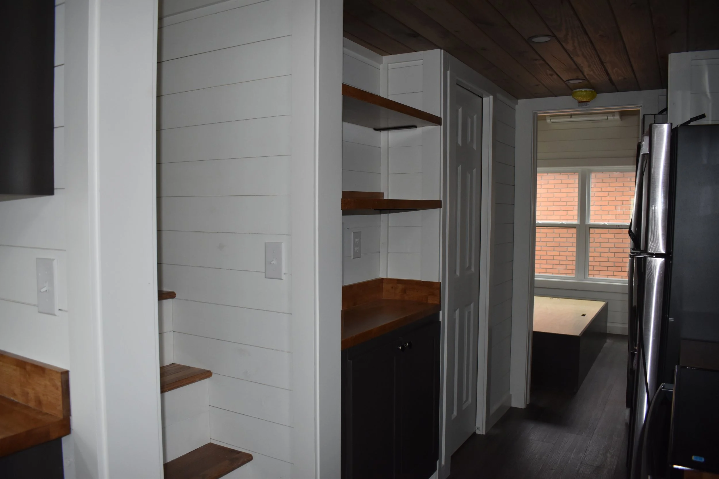 Interior view of a kitchen with white shiplap walls, wooden shelves, a small nook with a table by a window, and a stainless steel refrigerator.