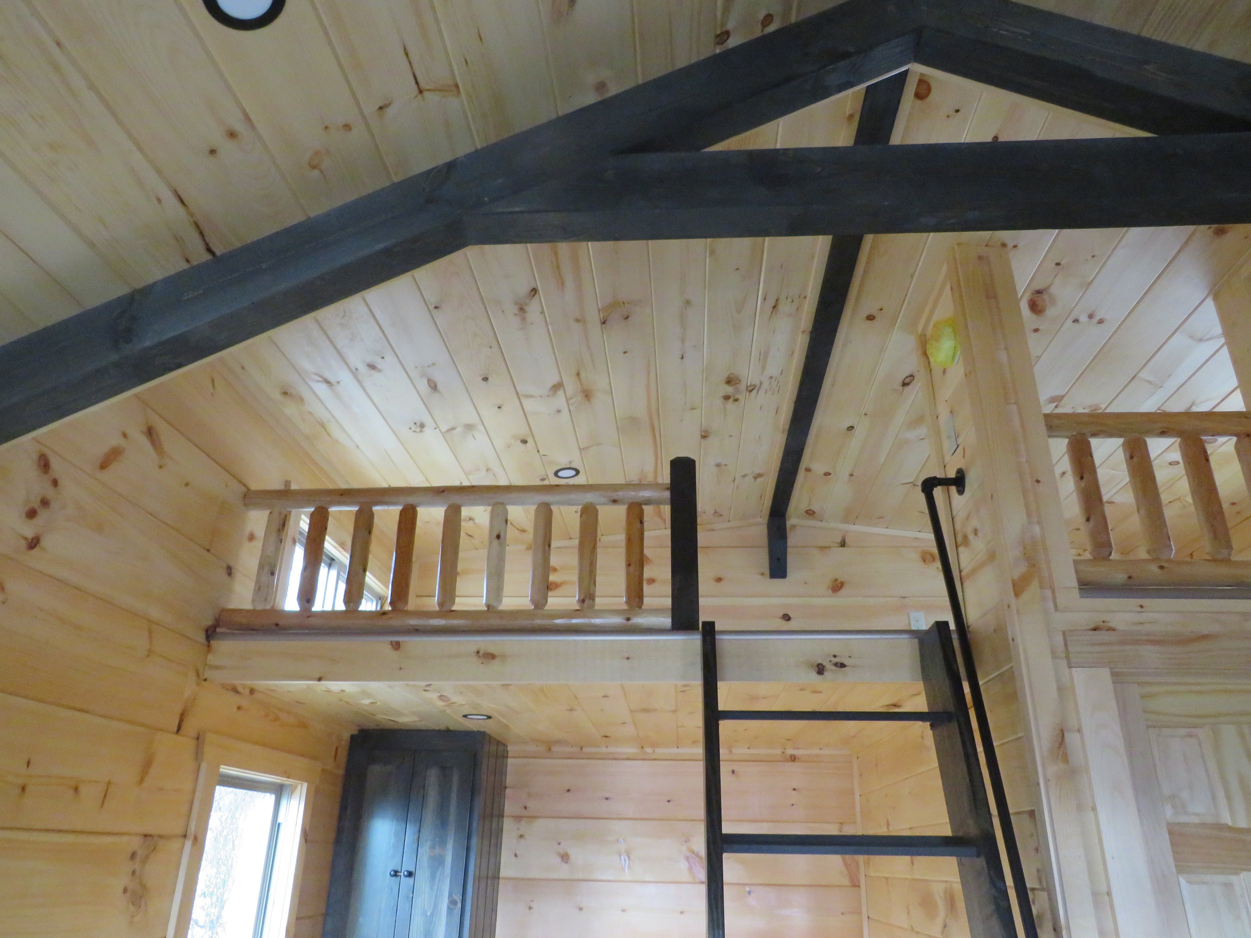 Interior view of a wooden room with a loft, staircase, and black railings, featuring natural wood paneling on the ceiling and walls.