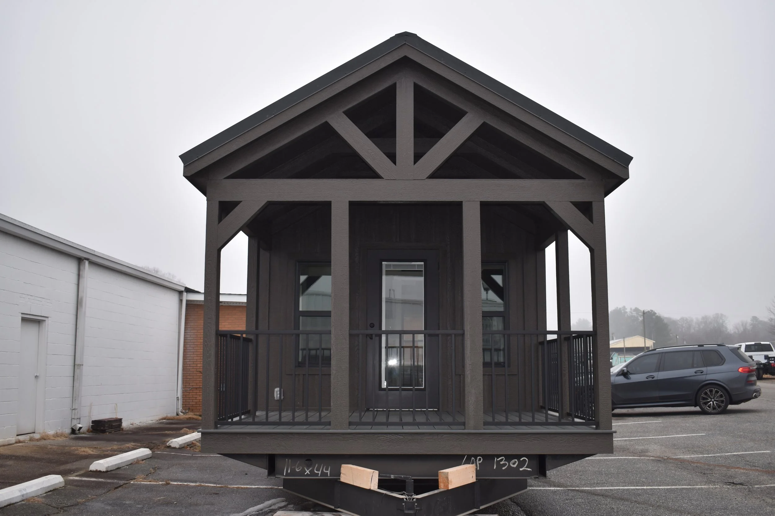 A small, dark gray wooden house with a porch and railing, parked in an empty lot on a cloudy day.